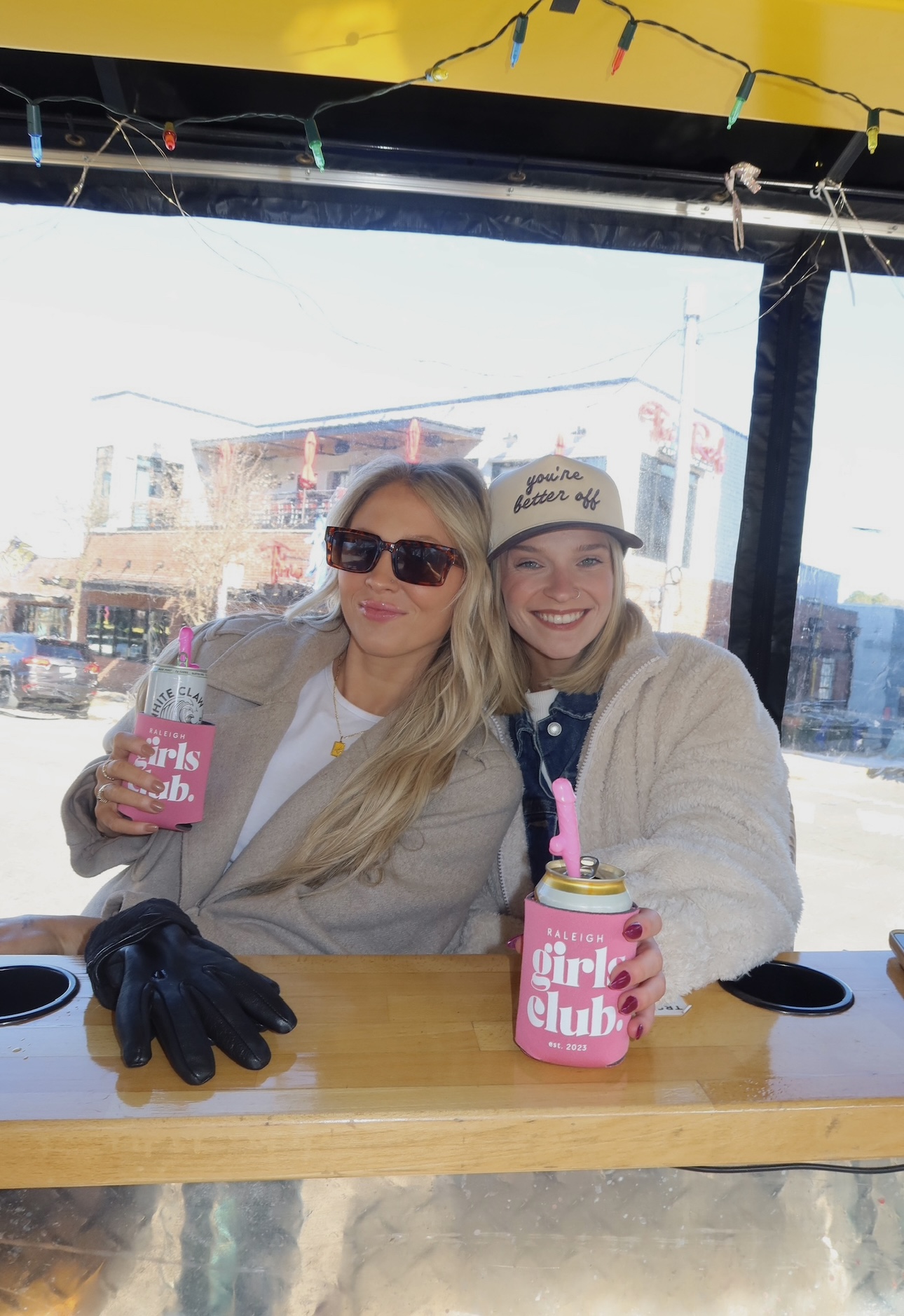 Two women sit at a table with pink drinks, smiling, in a bright cafe with large windows and colorful decorations.