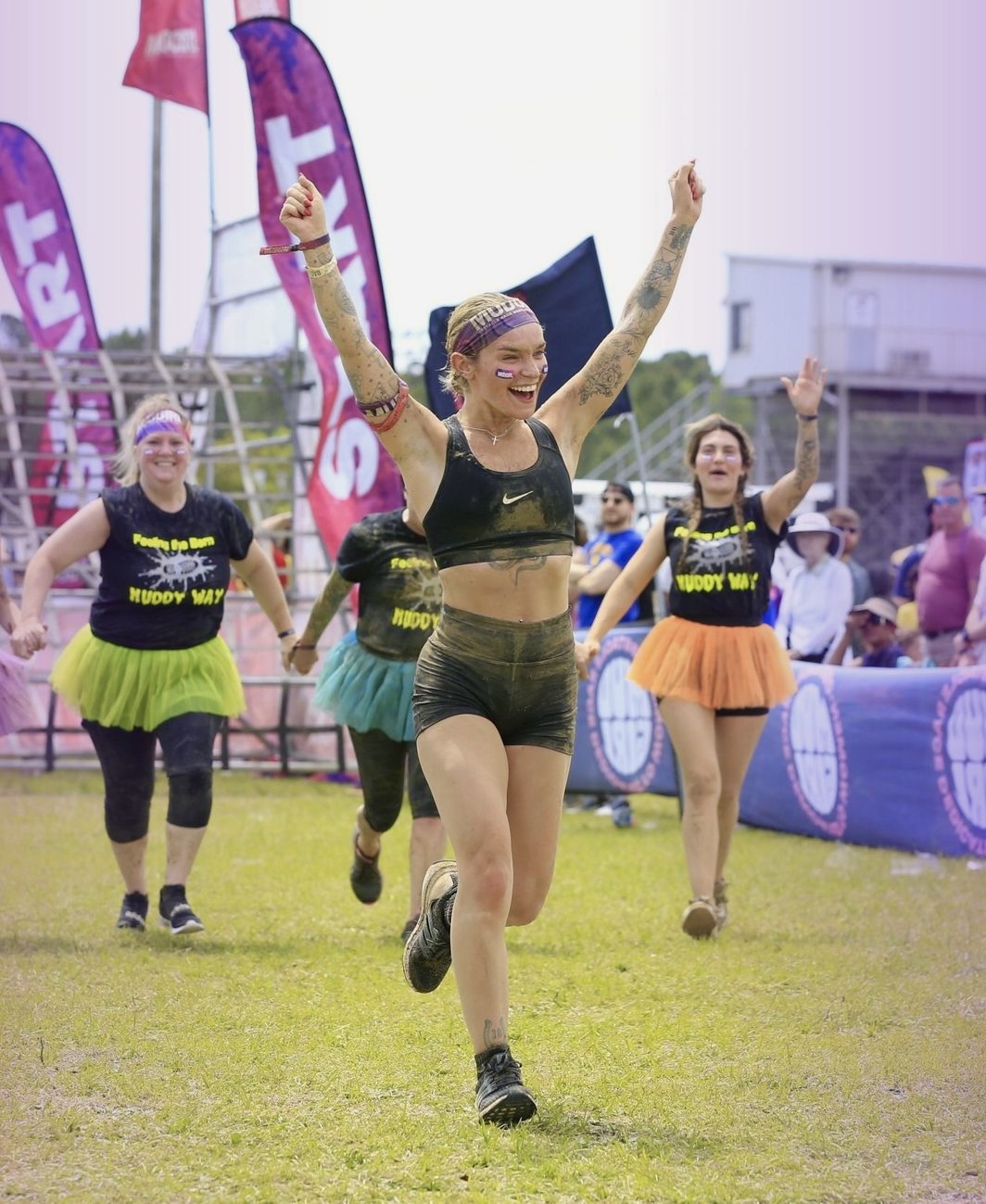 Group of women running on grass field, celebrating, wearing colorful athletic outfits, with flags and spectators in background.