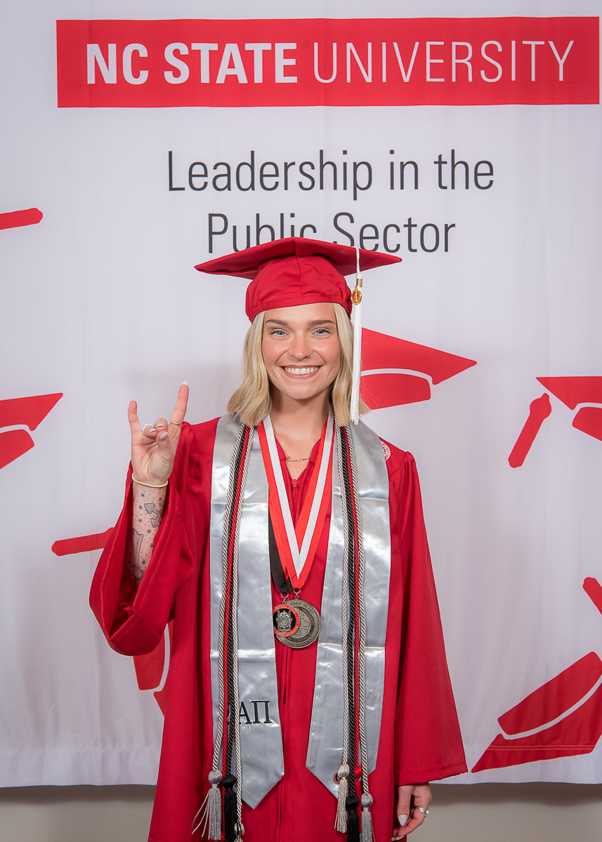 Young woman in graduation cap and gown smiling, holding up a hand with a rock sign, in front of a university banner.