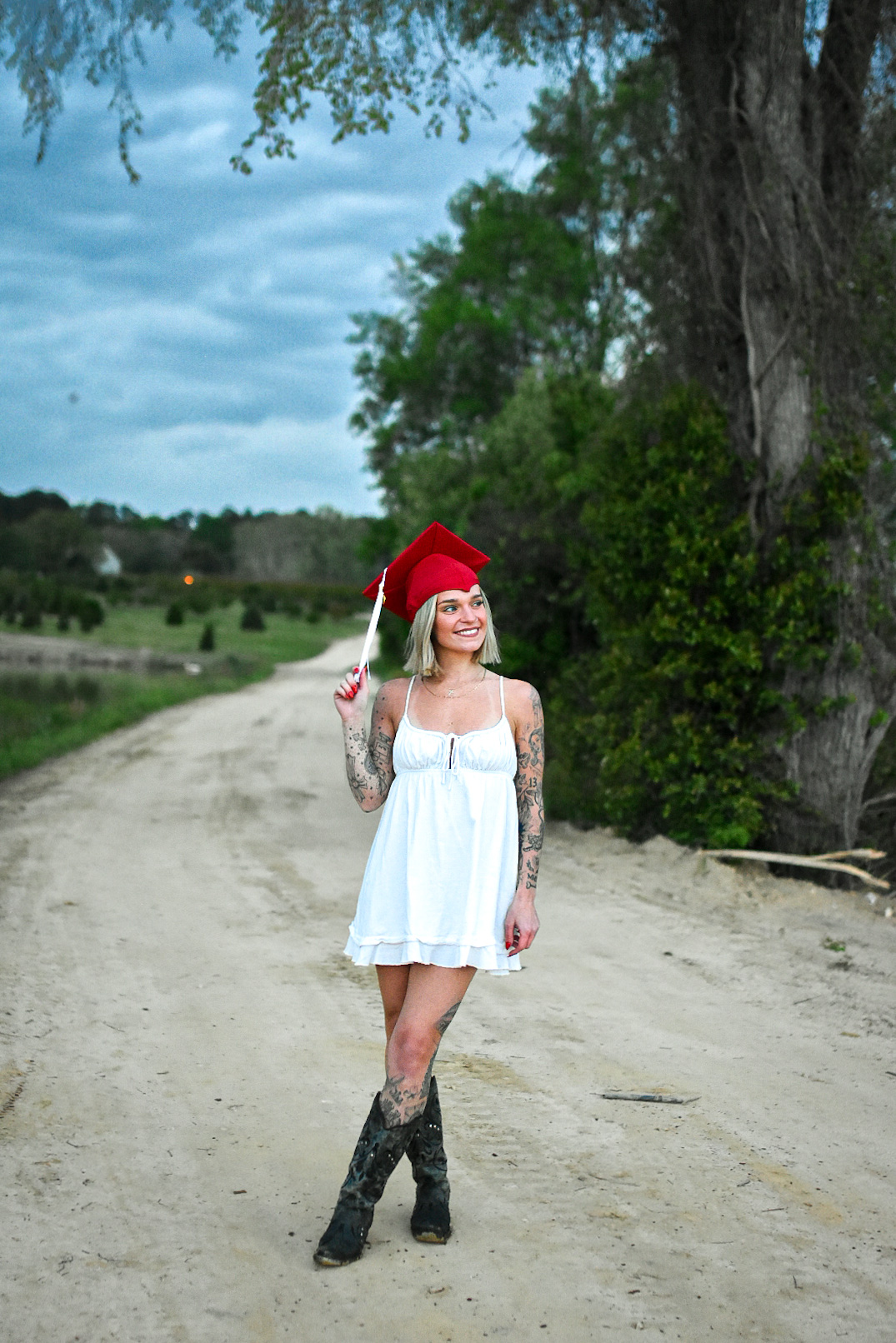 Young woman in a white dress and red graduation cap standing on a dirt path with trees and cloudy sky in background.
