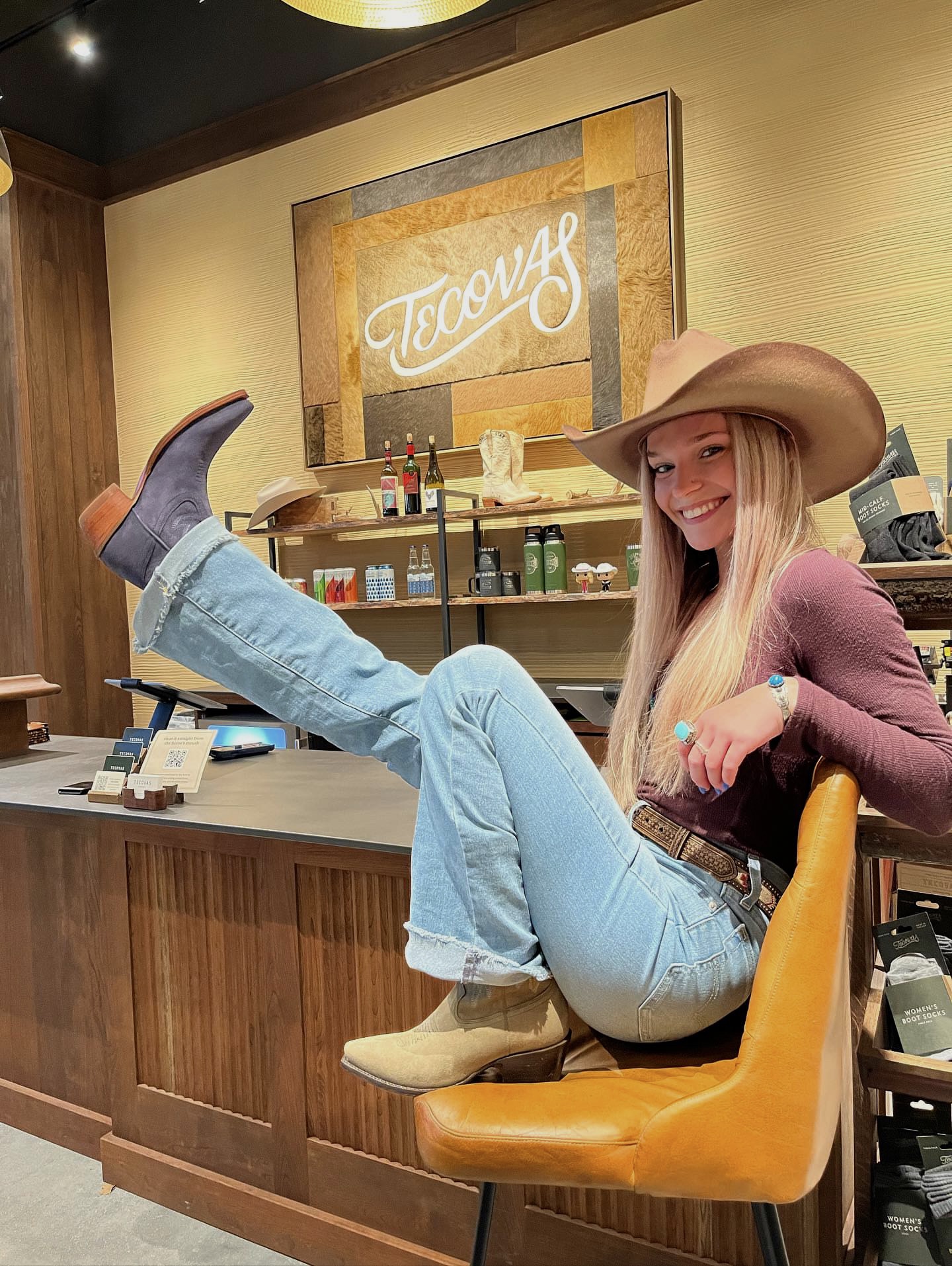 Young woman in cowboy hat and jeans sitting on a yellow chair in a store, smiling at the camera.