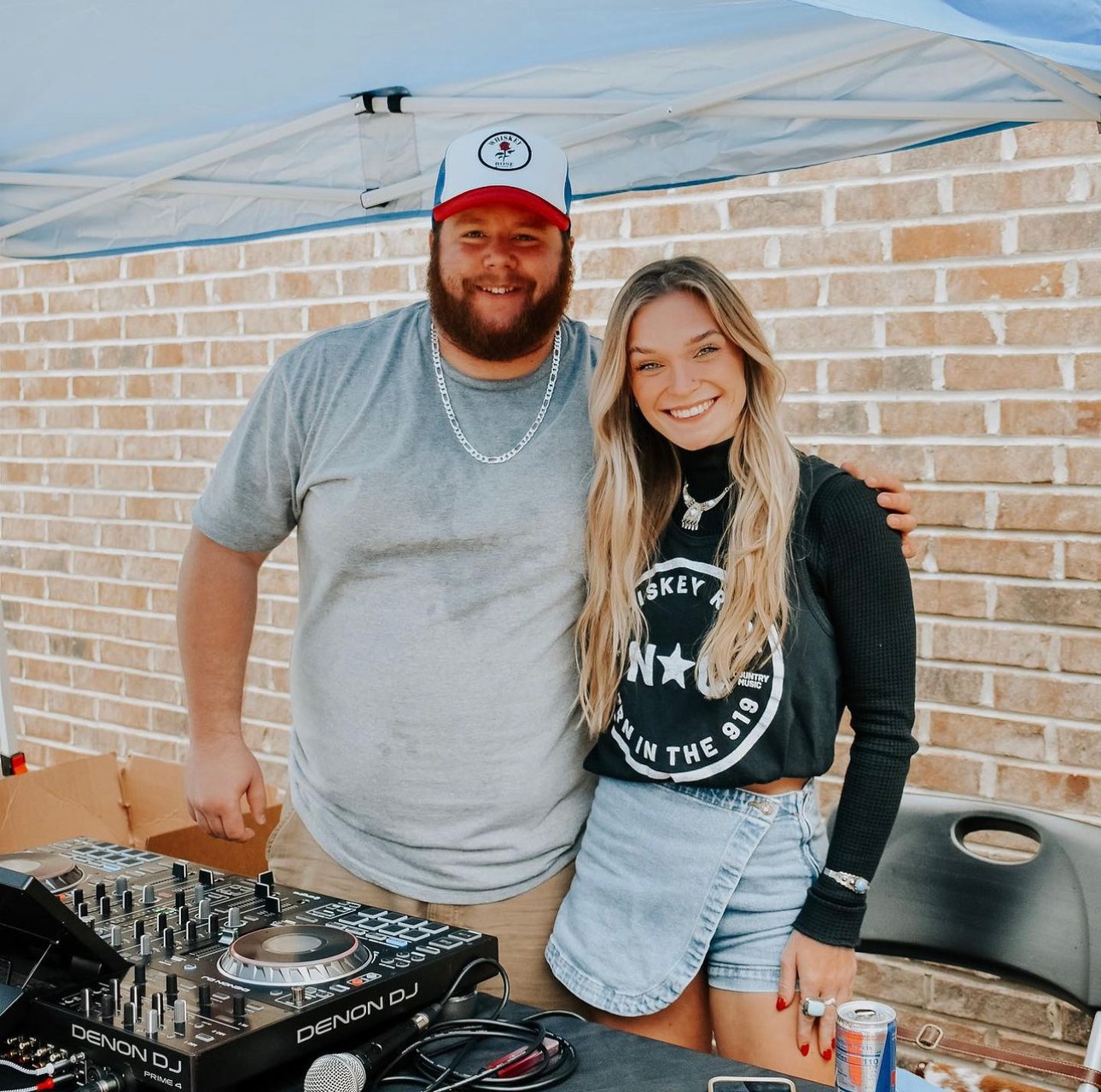 Two people smiling, standing outdoors under a canopy, with a brick wall behind them, a DJ setup in front.