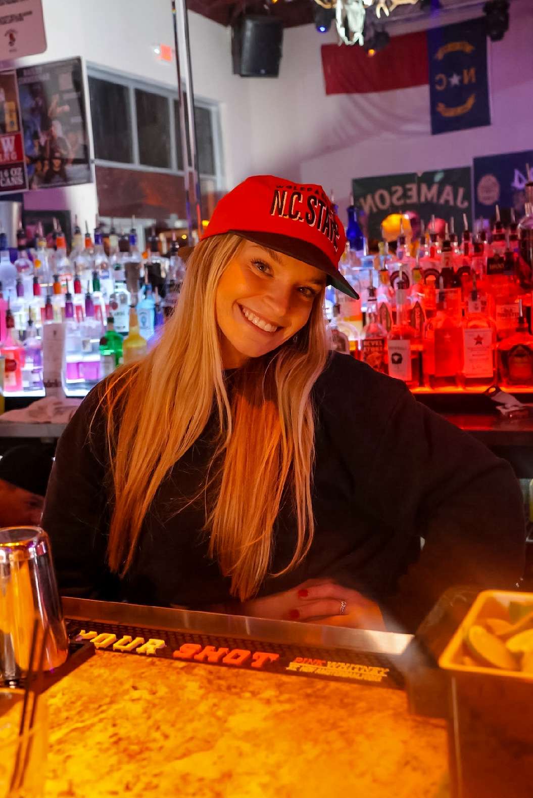 Woman with long hair wearing a red cap smiling at a bar with colorful bottles in background.
