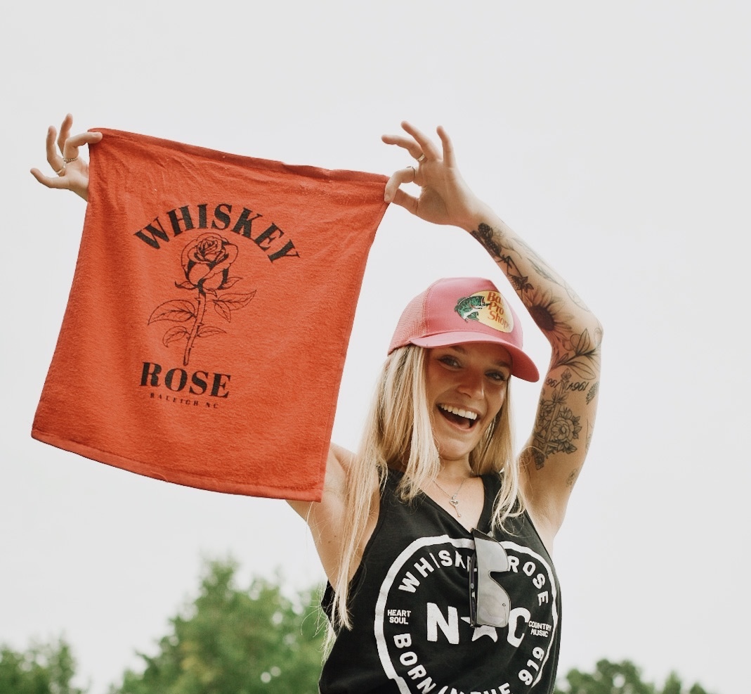 Woman holding a red cloth with 'Whiskey Rose' text, smiling, wearing a pink cap and black tank top, outdoors.