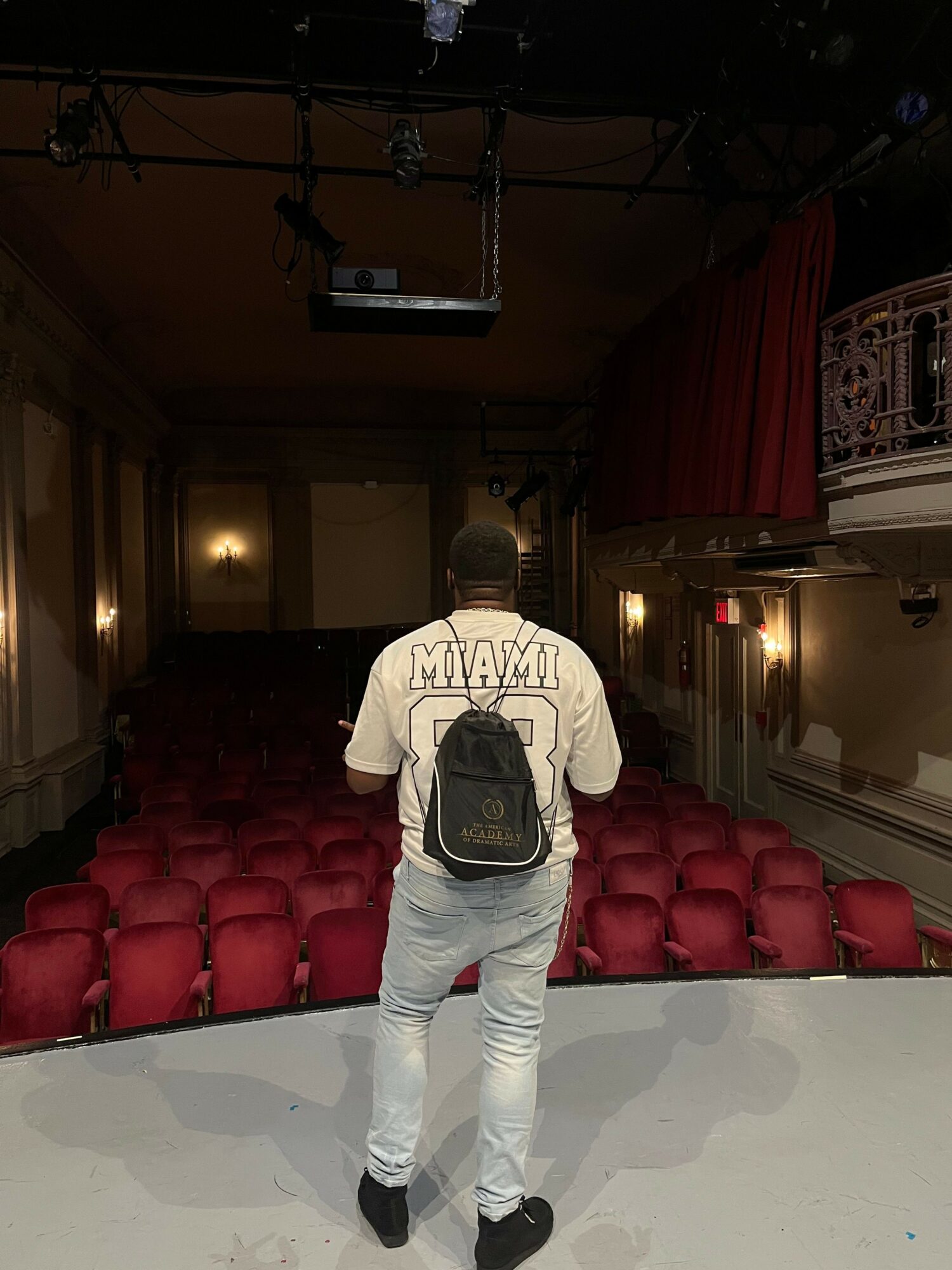 Person with a backpack taking a selfie in a room with red chairs and a white table, facing the camera.