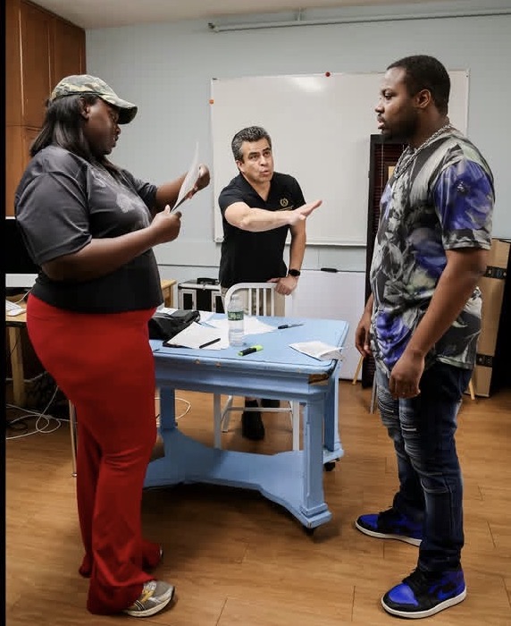 Three people in a room, one seated at a desk, two standing and talking, whiteboard in background, wooden floor.