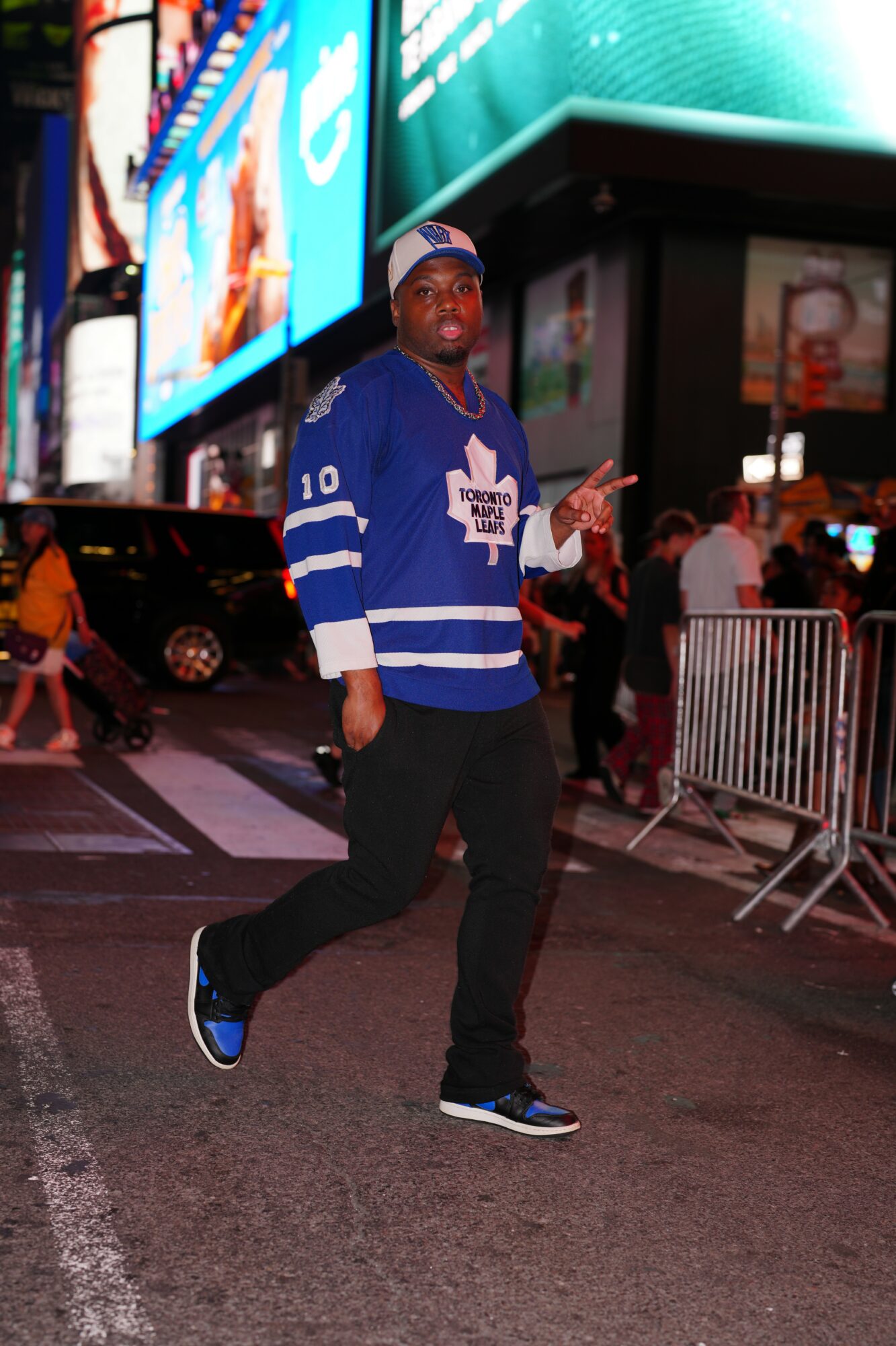 Man in Toronto Maple Leafs jersey walking in city at night with bright billboards and people around.