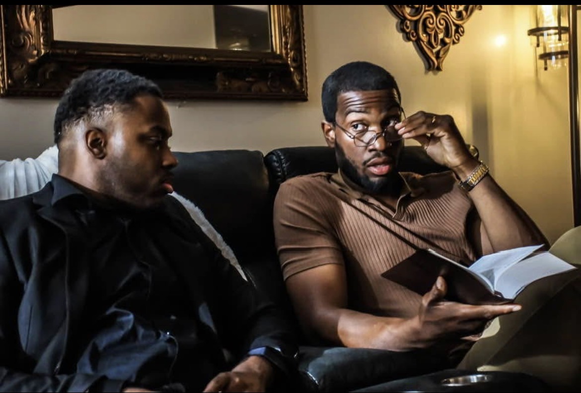 Two men sitting on a couch, one reading a book and the other adjusting glasses, in a dimly lit room.