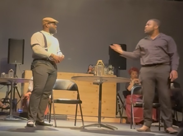 Two men are on stage, one gesturing while the other listens, with a woman sitting in the background. Stage setup includes a table and chairs.