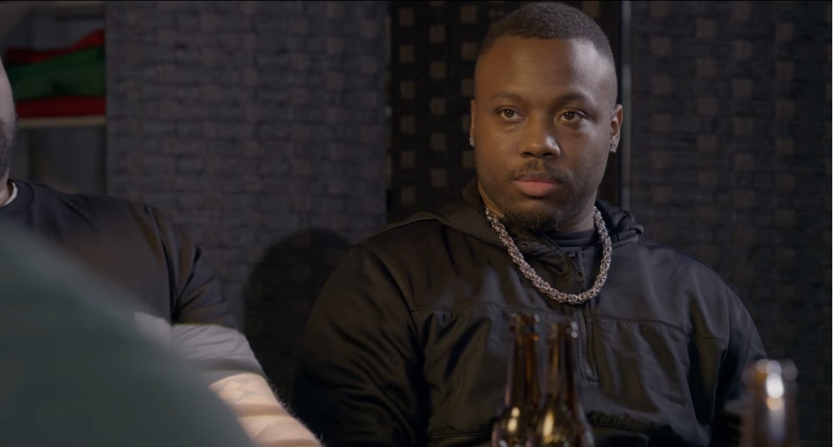 Man with short hair and chain necklace sitting indoors, dark background, serious expression.