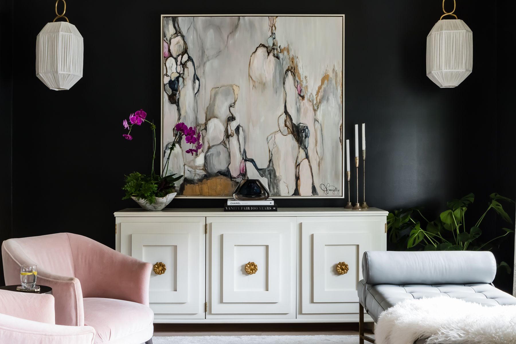 Living room with black wall, white sideboard, pink armchair, and abstract artwork, with hanging pendant lights and plants.