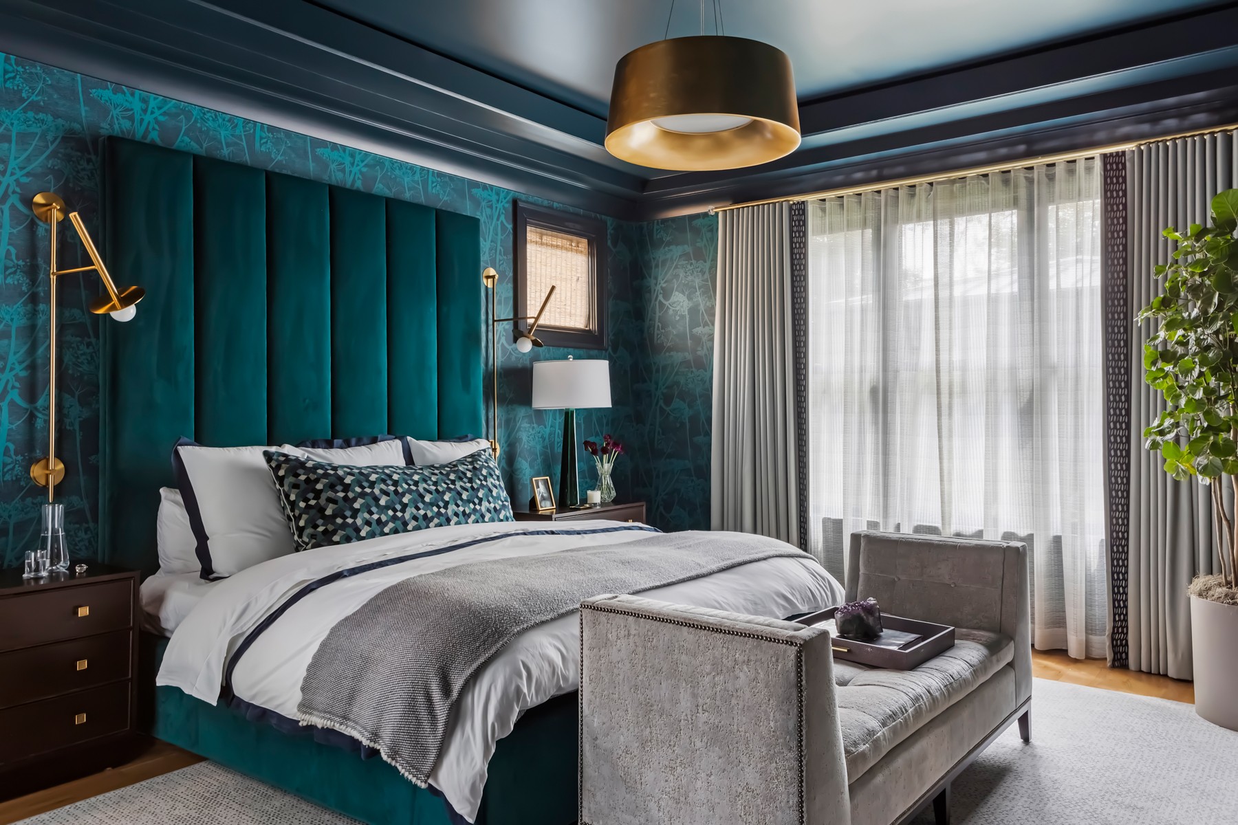 Bedroom with teal upholstered headboard, bed with gray and white bedding, window with curtains, and a bench at the foot of the bed.