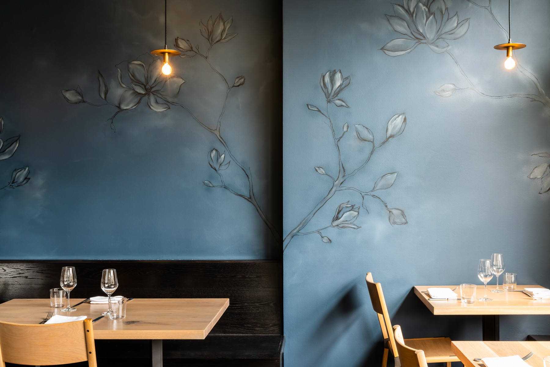 Two blue walls with floral patterns, two pendant lights, and tables set with glasses and napkins in a restaurant.
