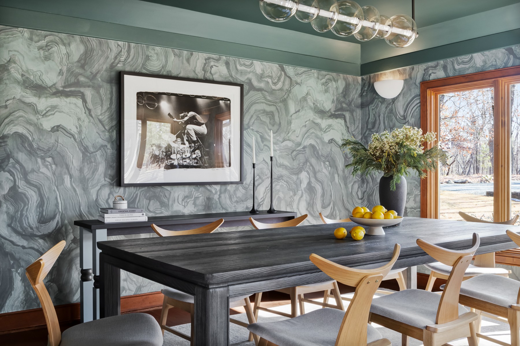 Dining room with a dark table, light chairs, a window, and a marble-patterned wall, with a vase of flowers and fruit on the table.