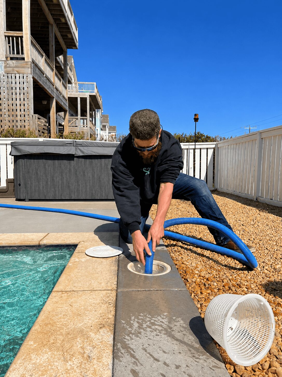 Person wearing sunglasses and dark clothing leaning over pool edge, handling a blue pool hose, with a white bucket nearby, outdoor setting.