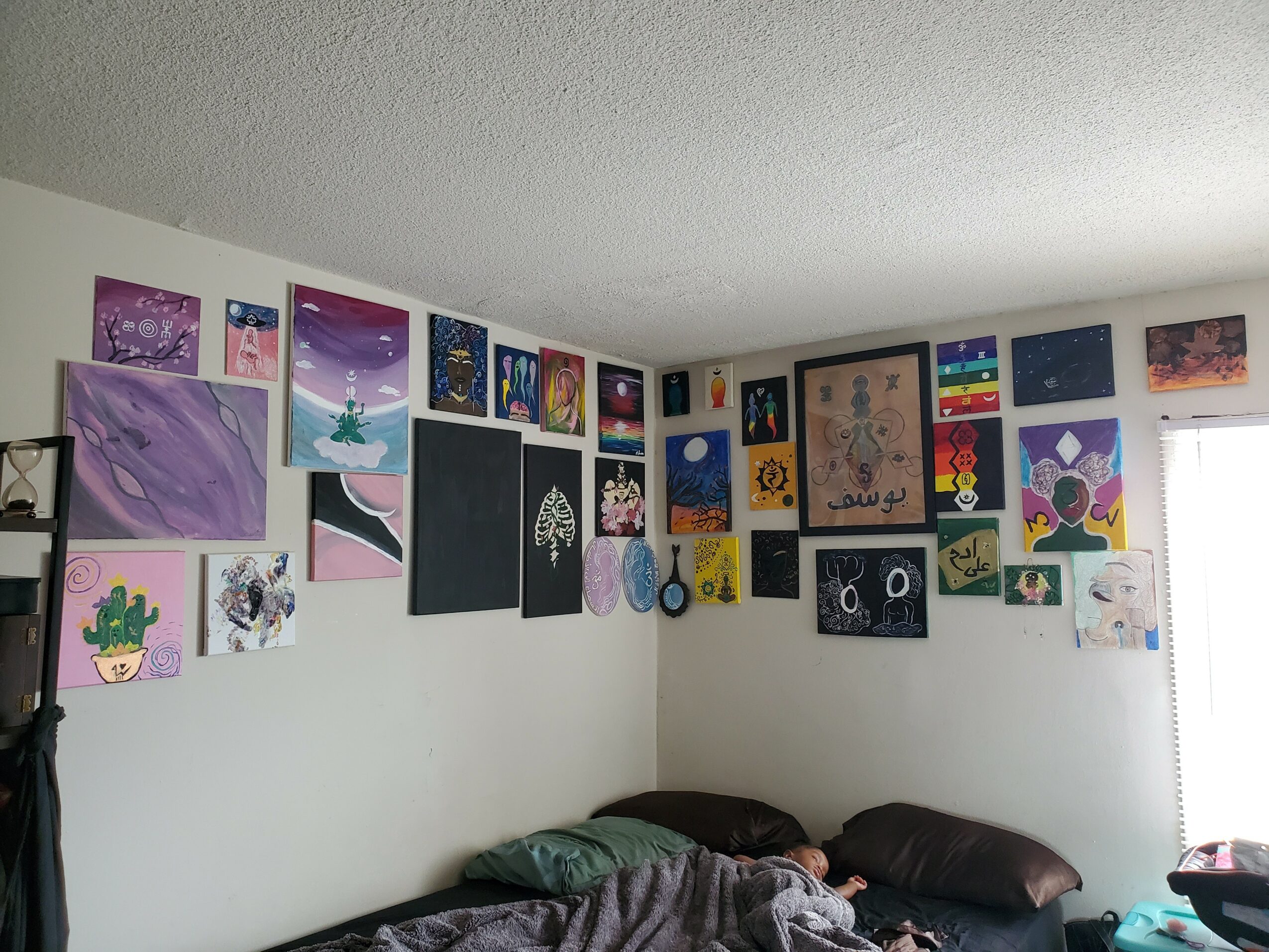 Gallery wall with colorful paintings and drawings above a bed with pillows, in a room with a window on the right.