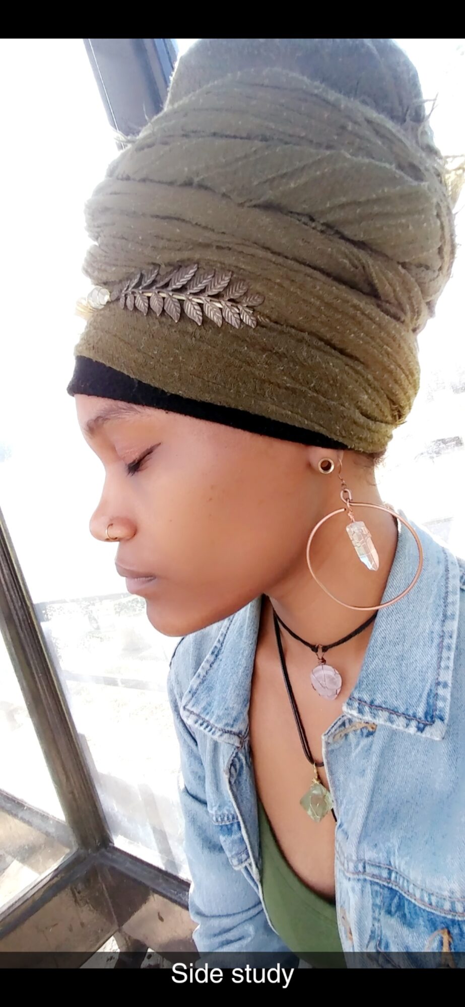 Side profile of a woman wearing a headwrap, earrings, layered necklaces, and a denim jacket, looking downward.