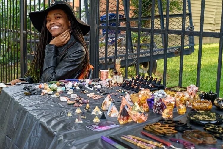Smiling woman wearing a hat sits behind a table with colorful gemstones and jewelry, outdoors near a black metal fence.