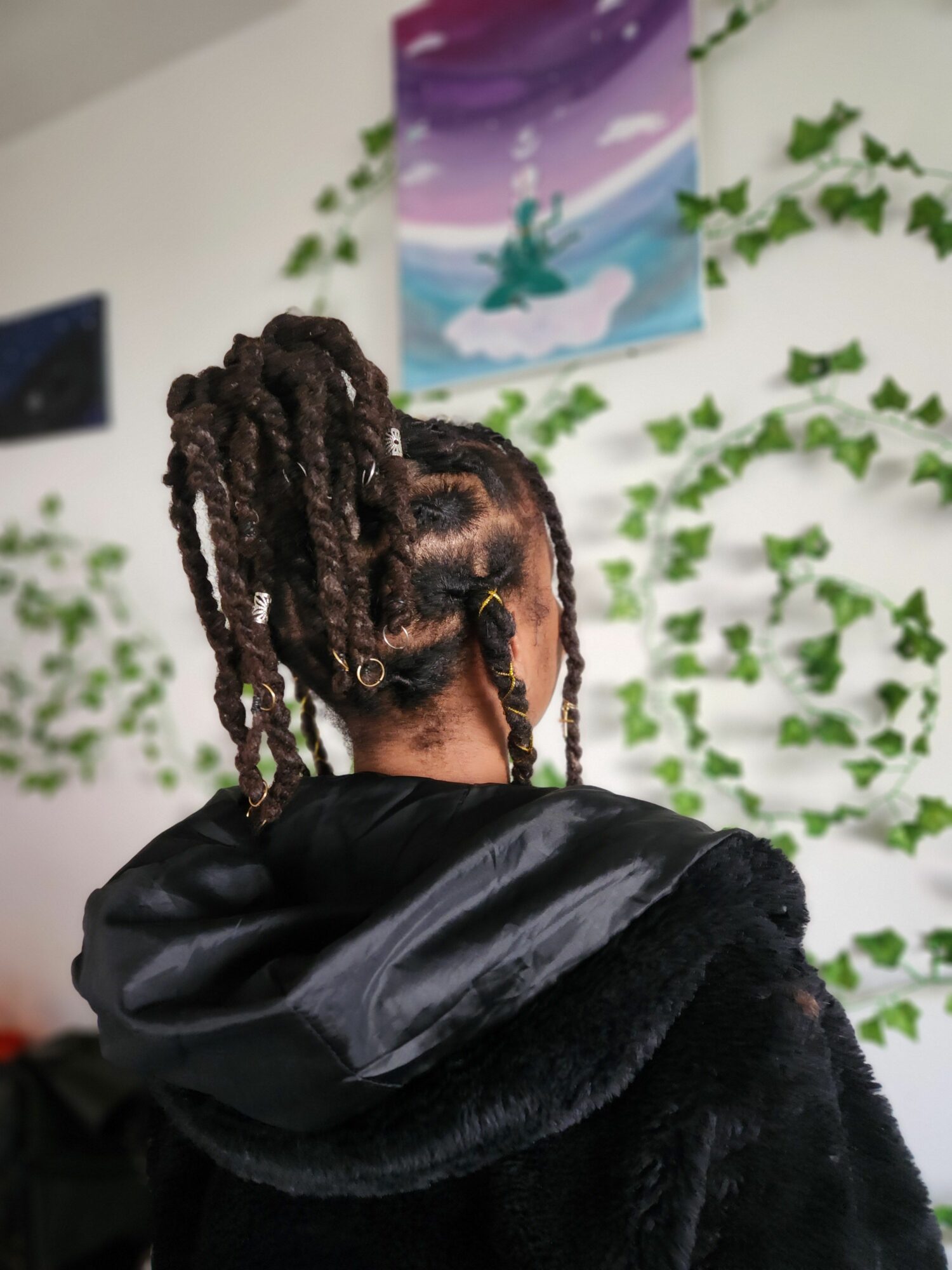 Person with dreadlocks wearing a black jacket, standing indoors with green plants and artwork in the background.