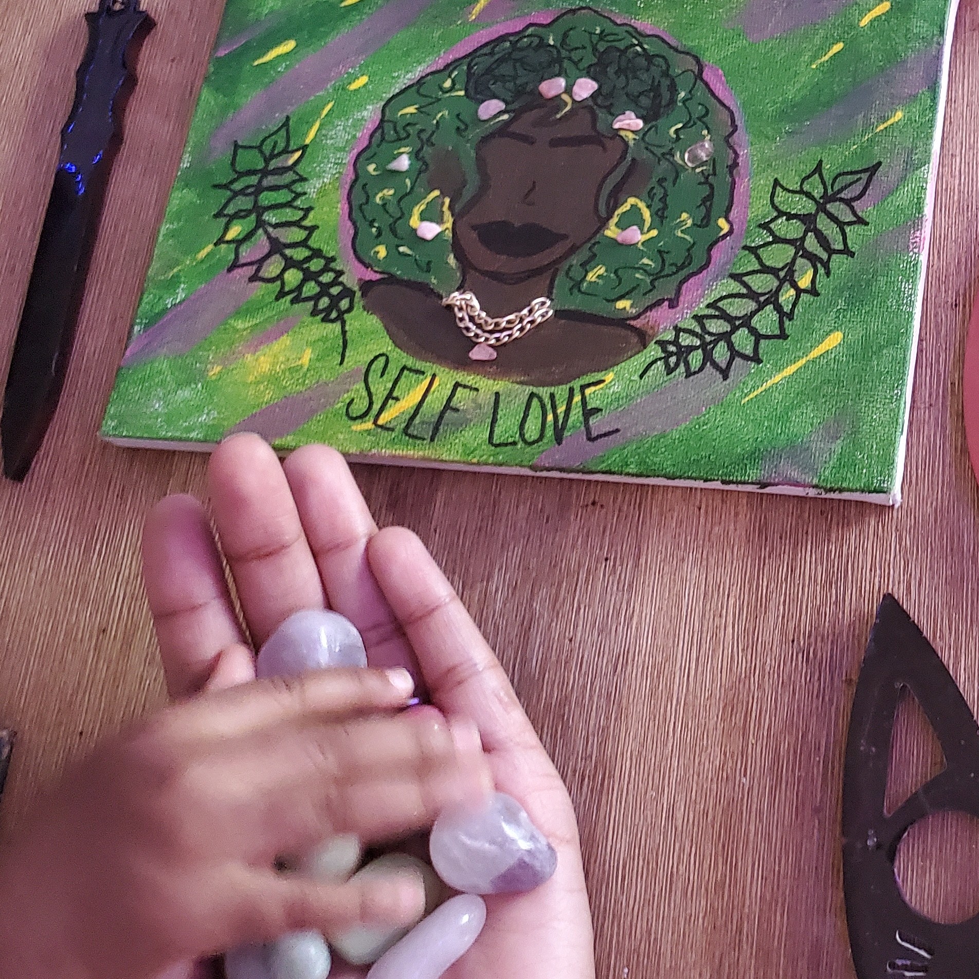 Painting of a person with green curly hair, surrounded by leaves, with the words 'Self Love' below, on a wooden surface.