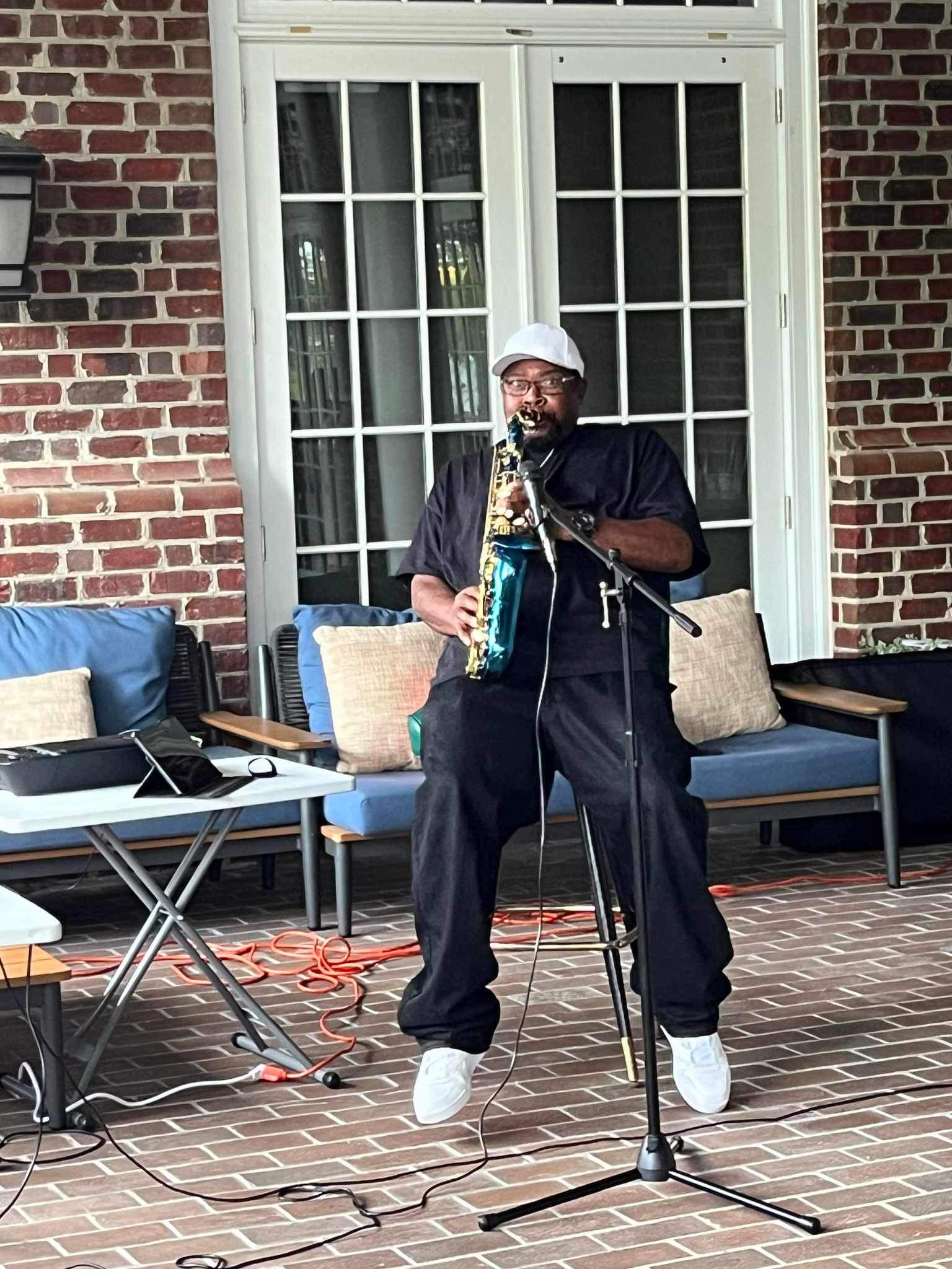 Man in dark clothing and white cap holding a microphone and a bottle, standing on a brick patio with outdoor furniture behind him.