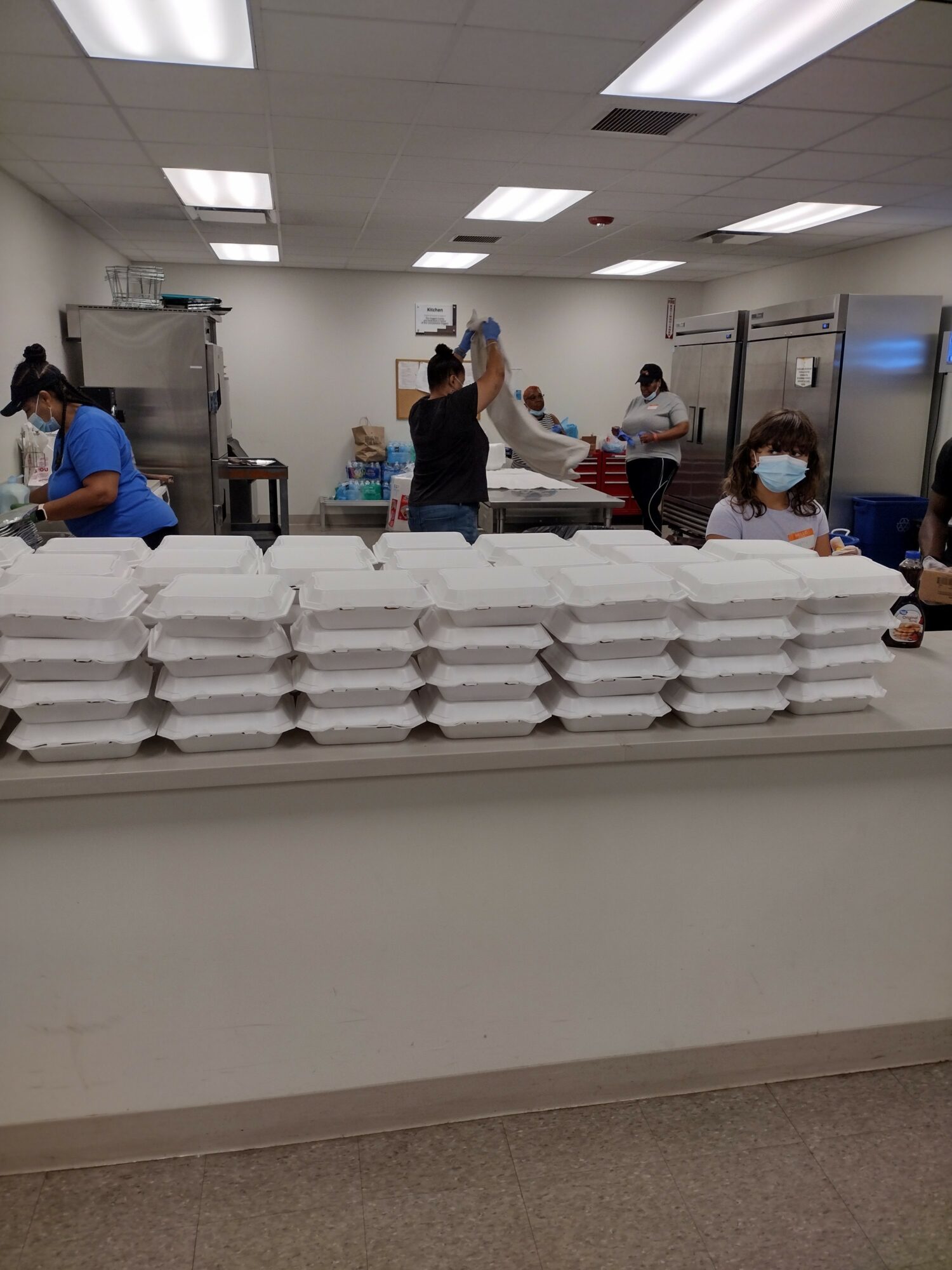 People in a room packing food containers, with stacked containers in the foreground, some wearing masks, fluorescent lighting overhead.