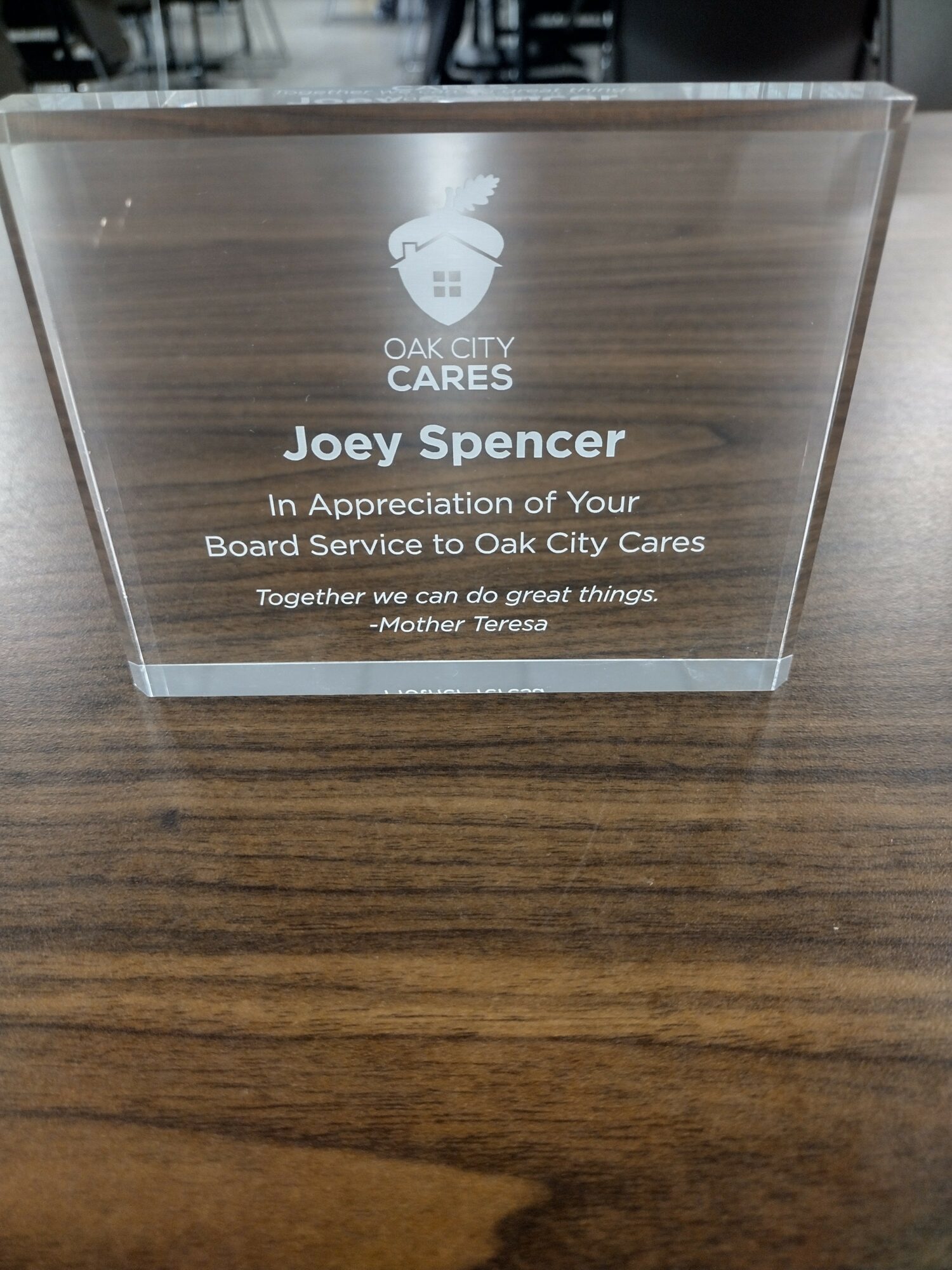 Clear glass plaque with engraved text and logo, placed on a wooden surface.
