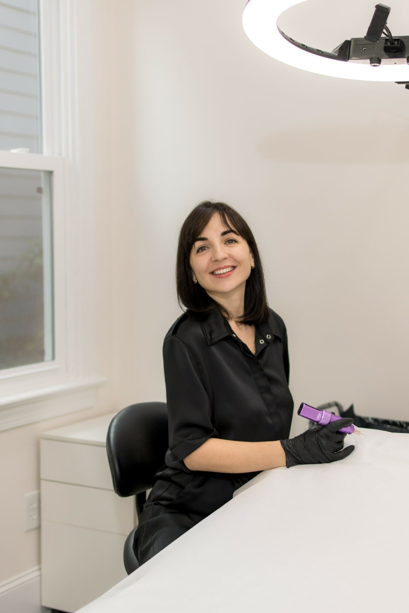 Young woman with dark hair smiling, wearing black gloves and shirt, sitting at a white table in a bright room.