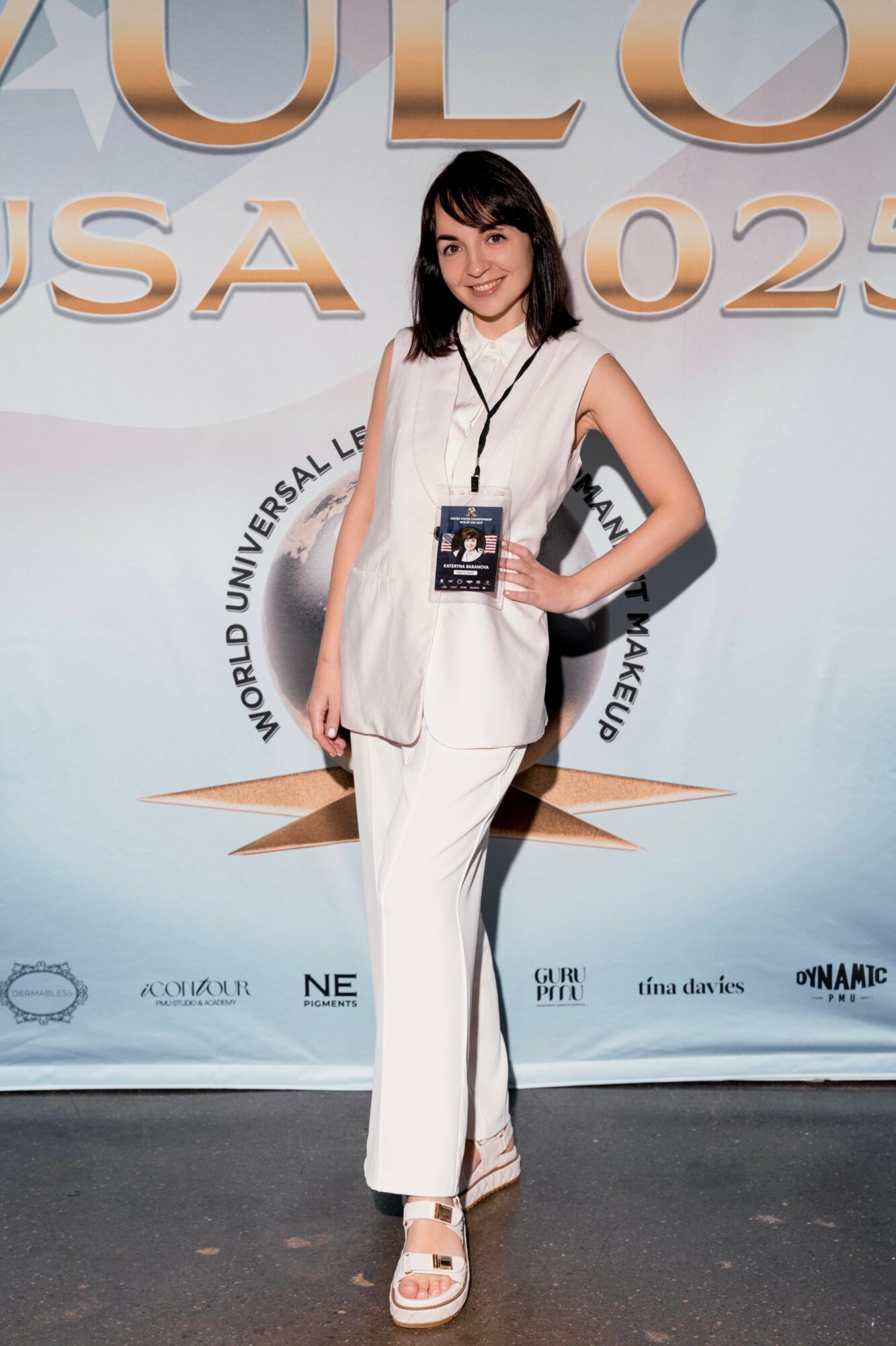 Young woman in white outfit holding a badge, standing in front of a backdrop with large text and logos.