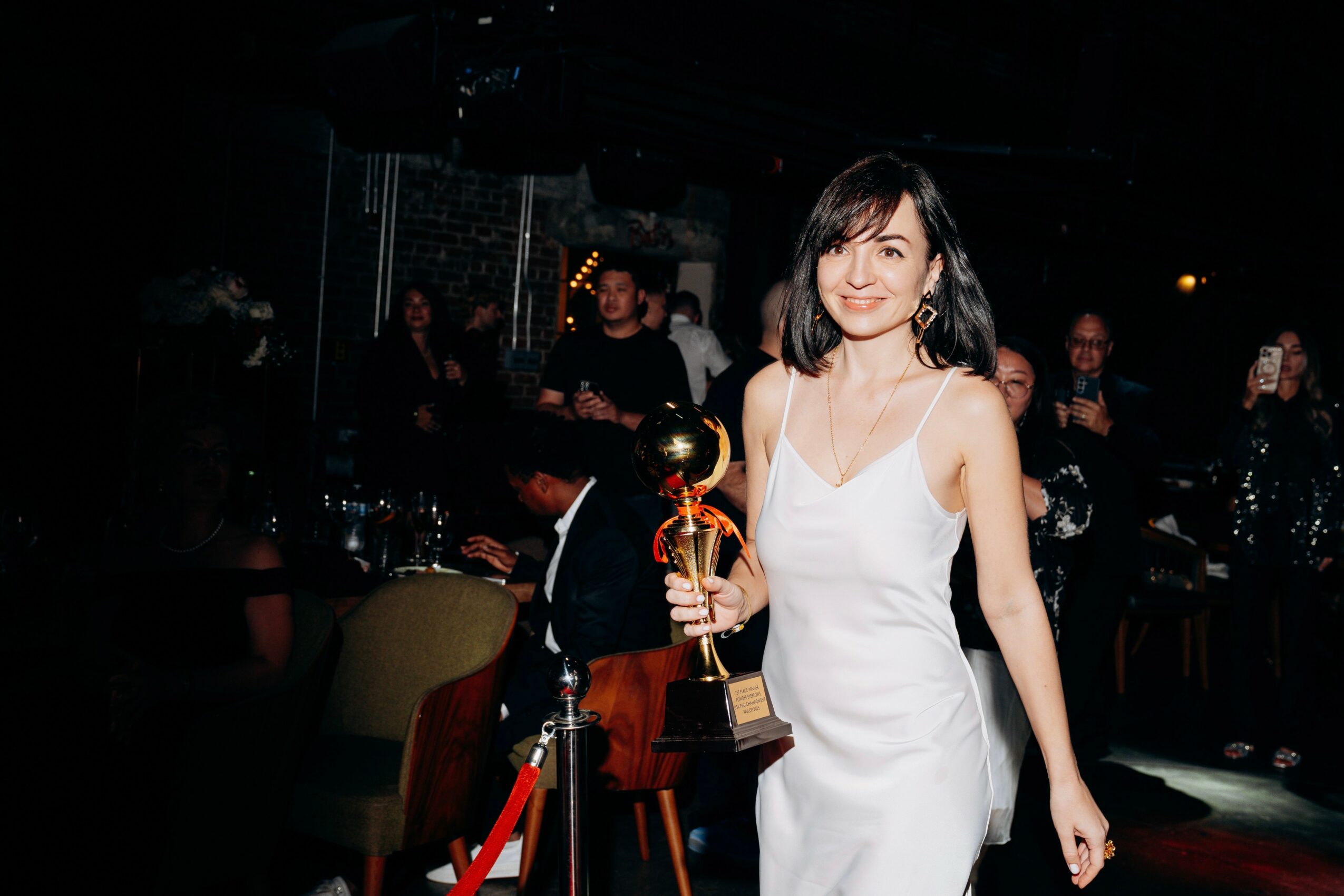 Young woman in white dress holding a trophy at an indoor event with people in the background.