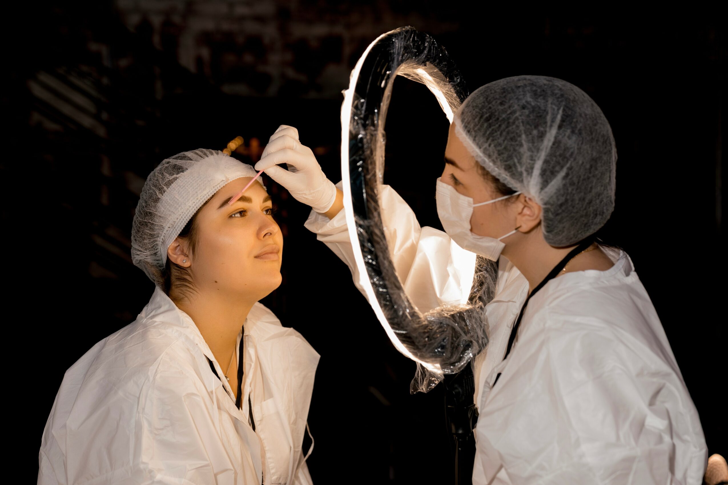 Medical professional examines a woman's face using a ring light, both wearing protective gear, in a clinical setting.