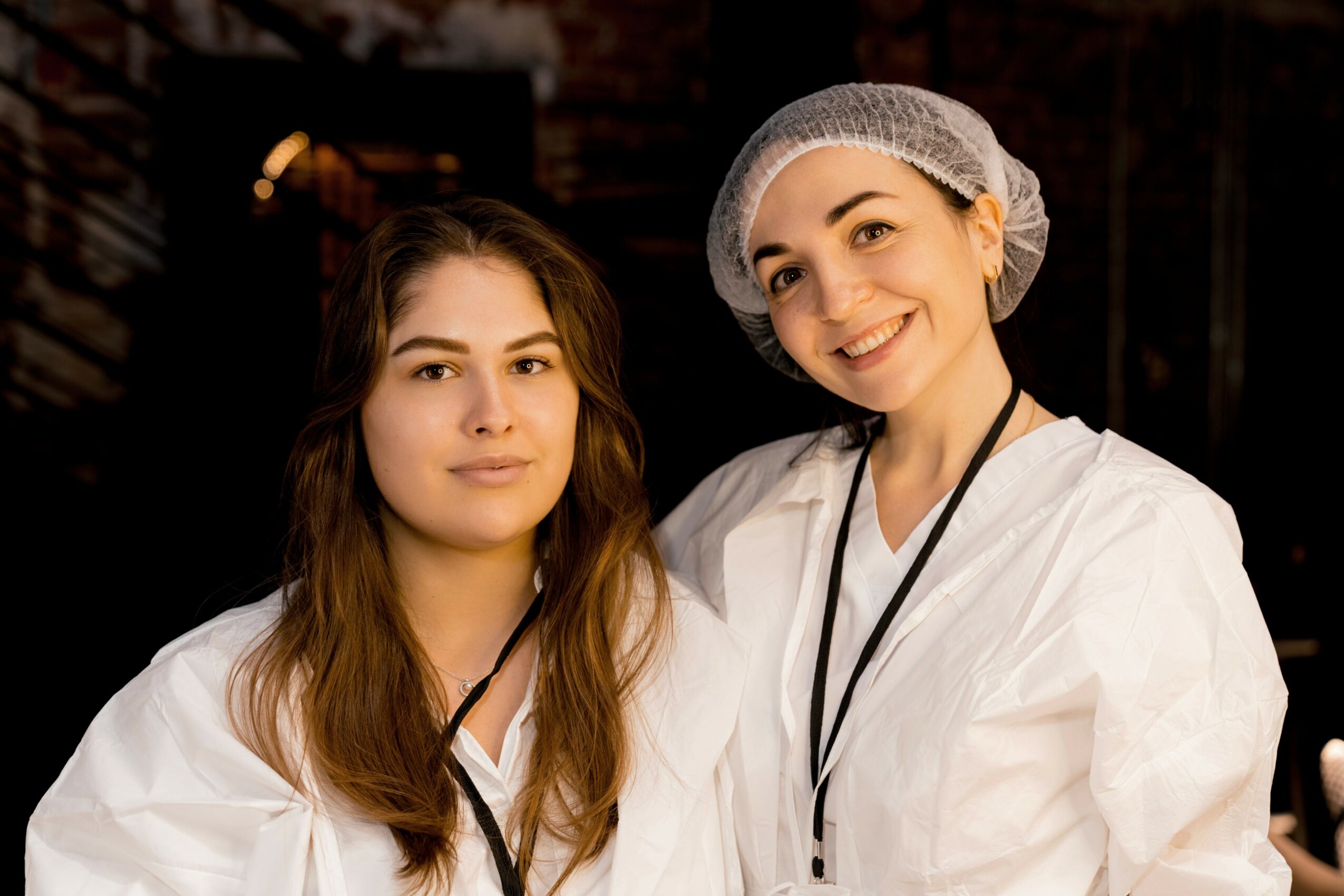 Two women in white uniforms smiling indoors, one wearing a hairnet, the other with long hair, in a professional setting.