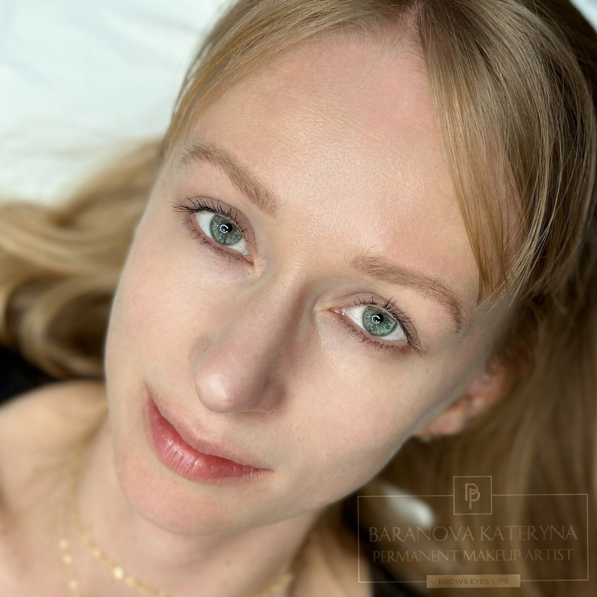 Close-up of a woman with blonde hair and blue eyes, smiling softly, lying down, with natural makeup and a gentle expression.