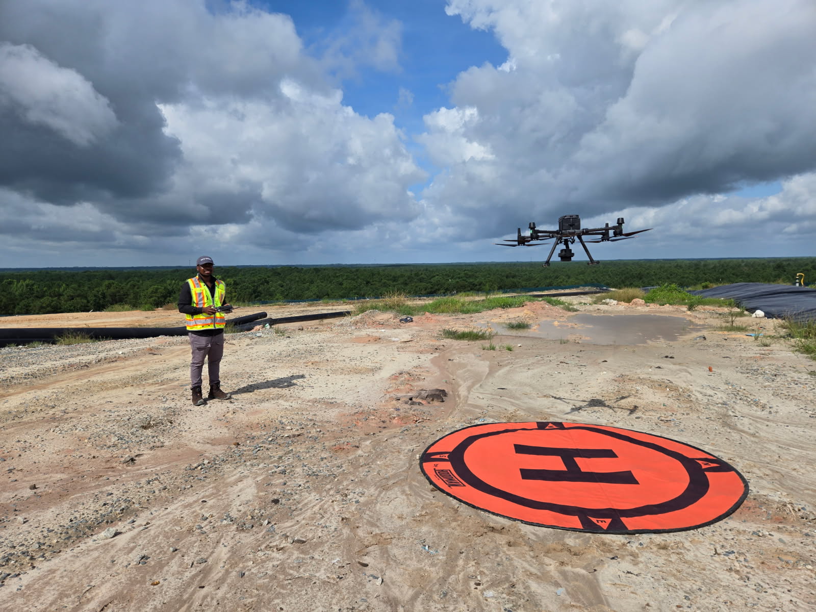 Person in safety vest operating a drone on a construction site with a large helipad symbol, cloudy sky in background.