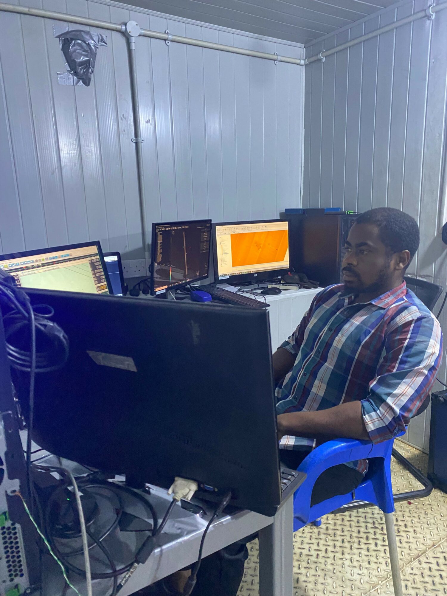 Person sitting at a desk with multiple computer monitors and a laptop, in a room with metal walls.