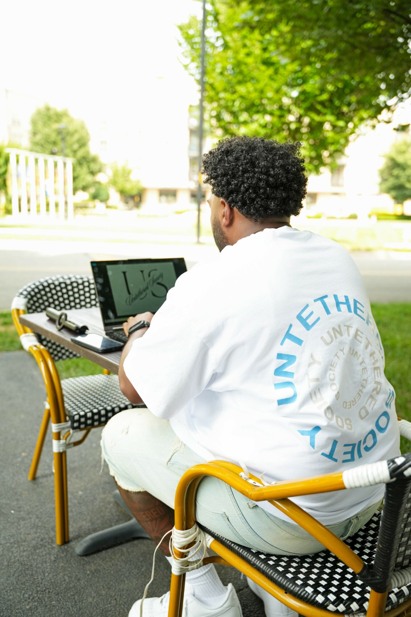 Person with curly hair sitting outdoors at a table, working on a laptop, with trees and bright sky in background.