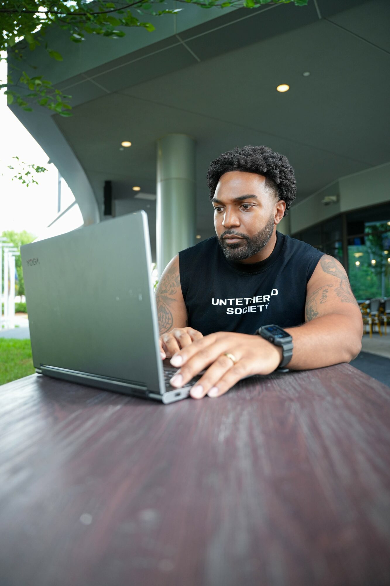 Man with curly hair and a beard working on a laptop outdoors under a covered area, wearing a sleeveless shirt and watch.