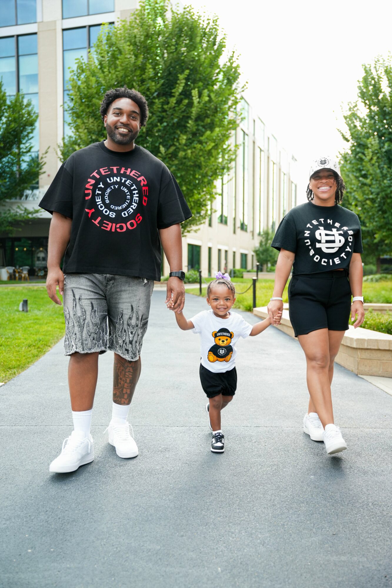 Family of three walking outdoors on a paved path, holding hands, with trees and modern buildings in the background.