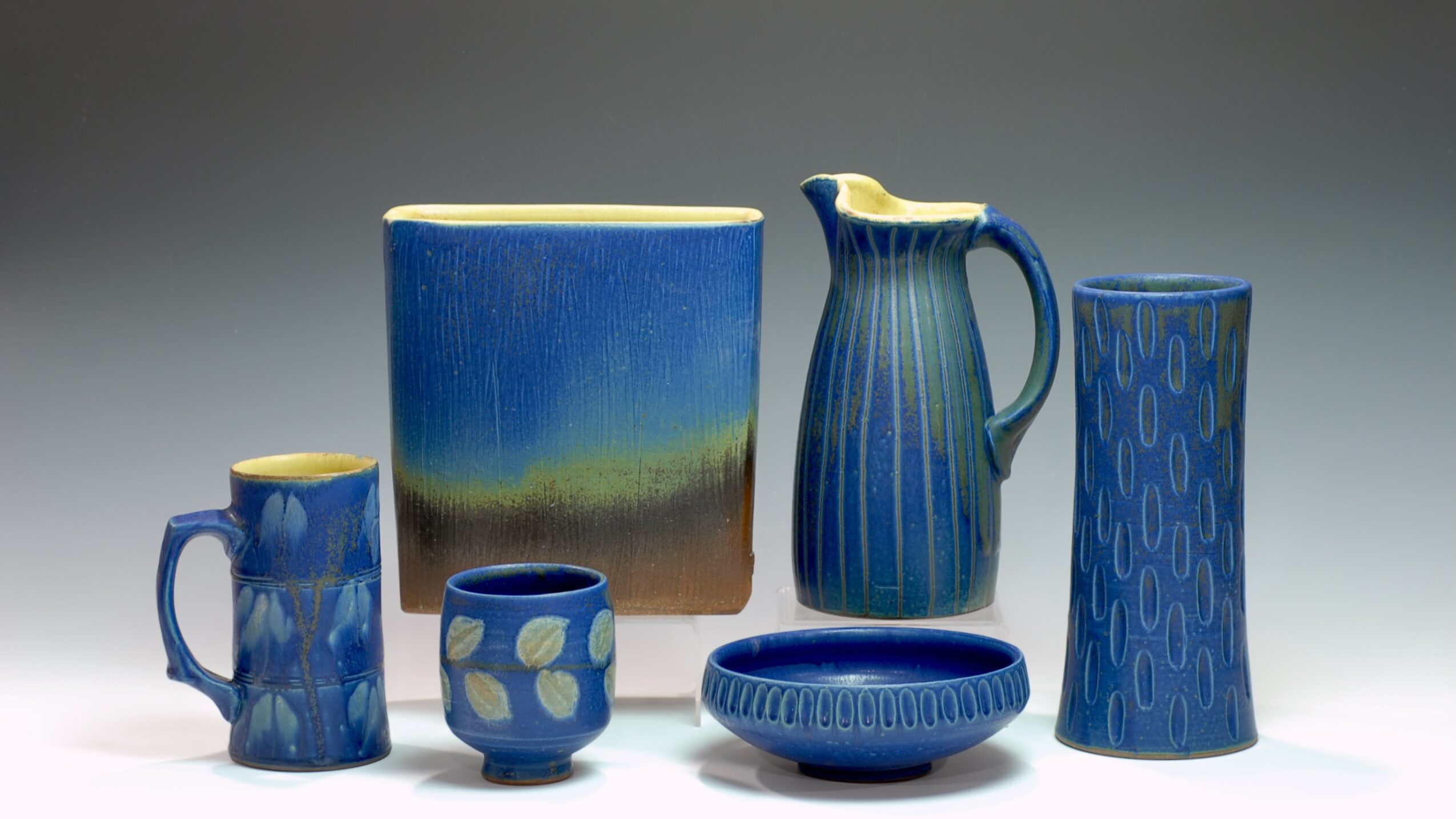Collection of blue ceramic vessels including a mug, small cup, bowl, pitcher, and tall vase, on a neutral background.