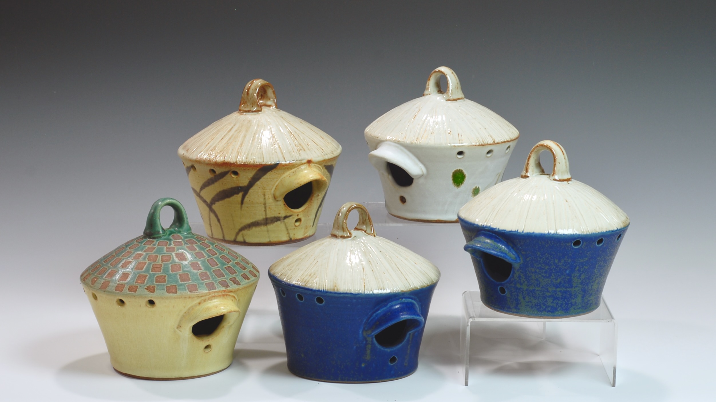 Five ceramic teapots with lids, in various colors and patterns, displayed on a white surface against a gray background.