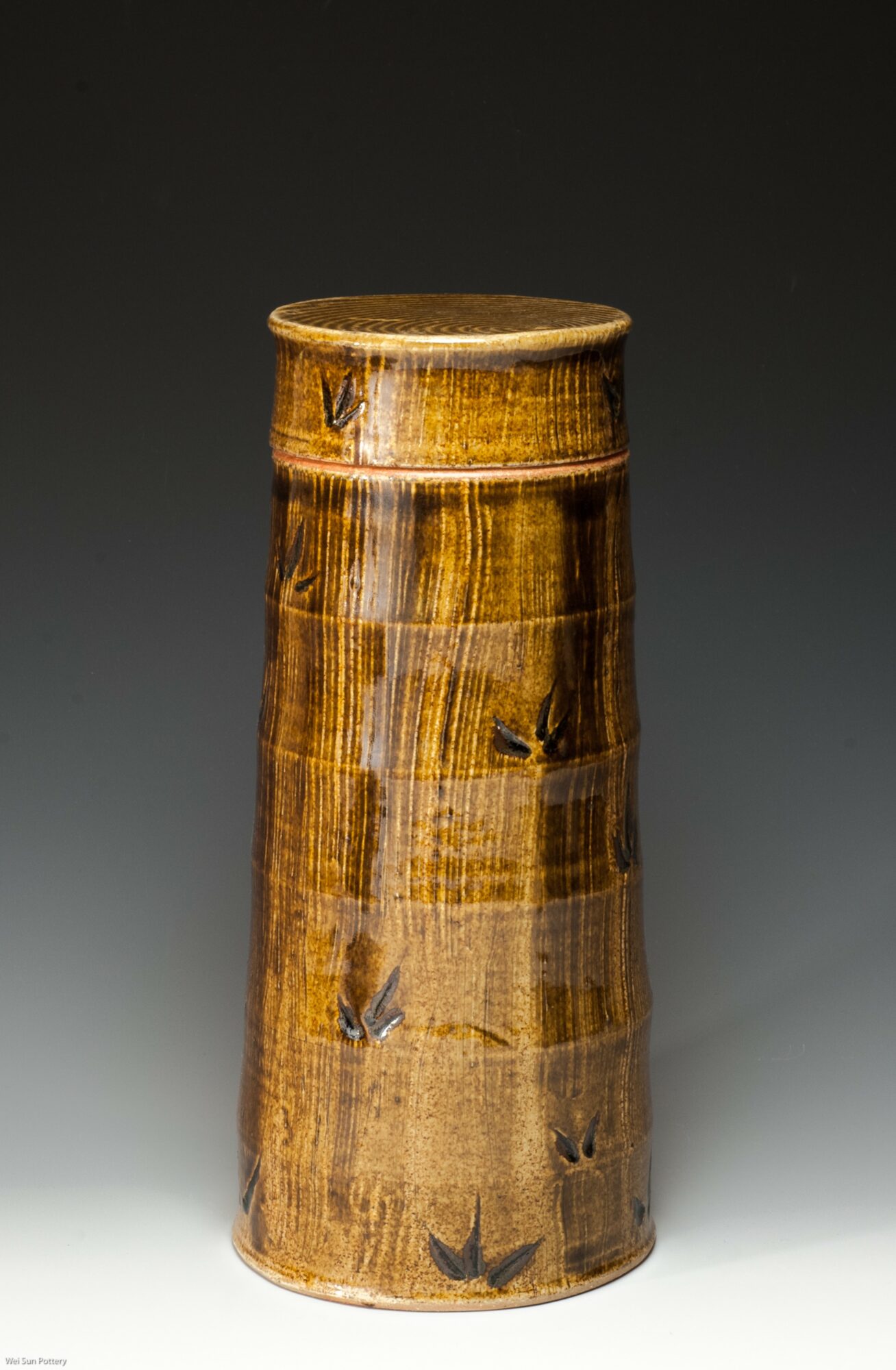 Tall, cylindrical ceramic vase with a glossy brown glaze and subtle decorative patterns, set against a dark background.