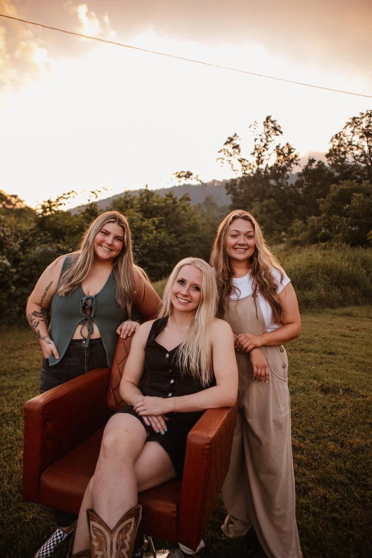 Three women outdoors, one sitting in a chair and two standing behind, smiling, with trees and sky in background.