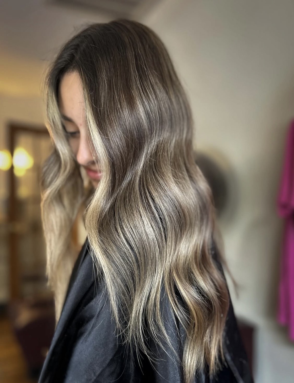 Side view of a woman with long, wavy, ombre hair, indoors with blurred background.