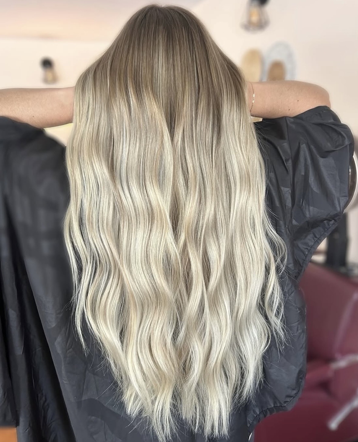 Back view of a person with long, wavy, blonde hair, wearing a dark top, in a room with light-colored walls.