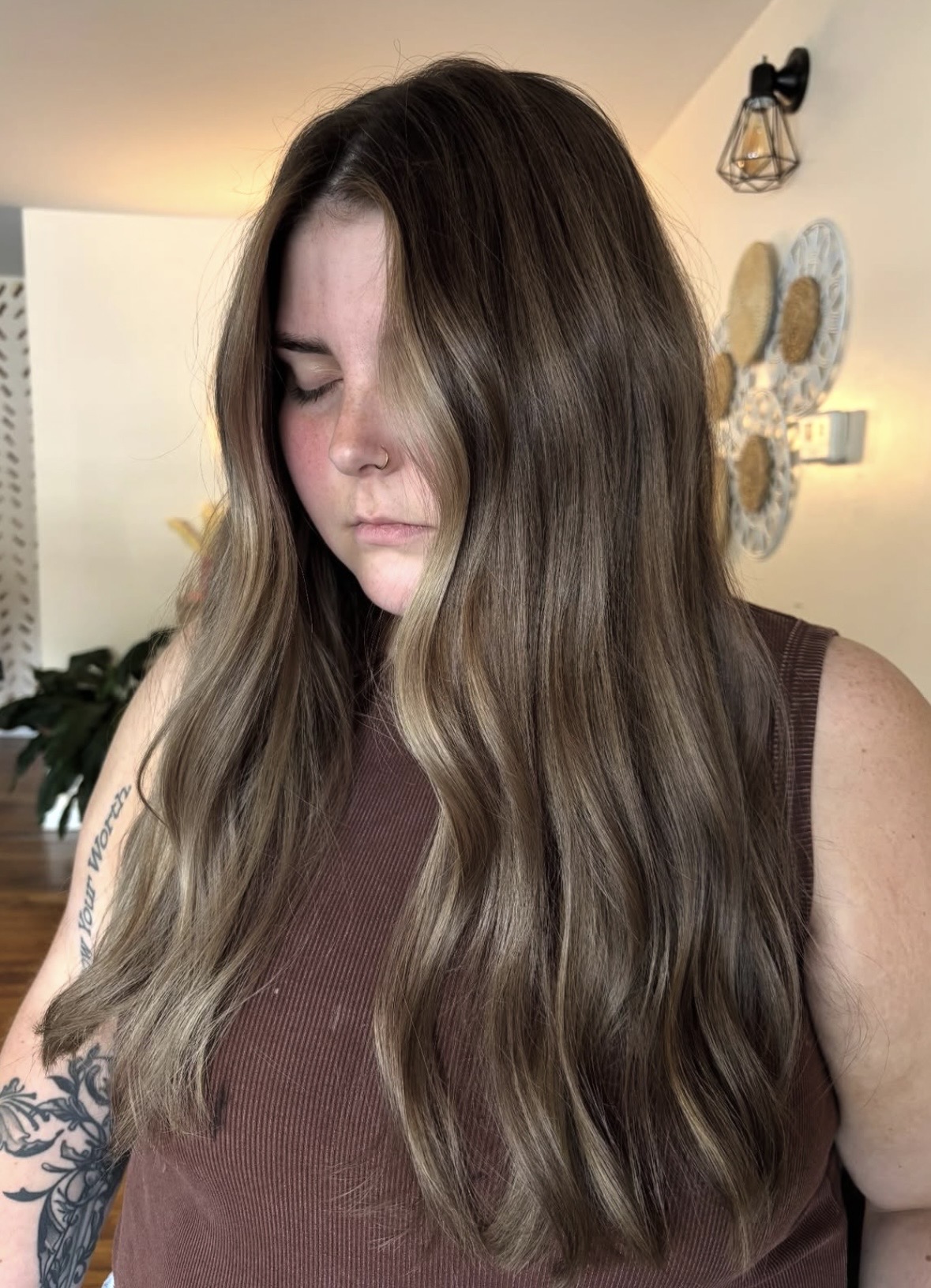 Young woman with long, wavy brown hair looking down, wearing a sleeveless brown top, in a room with wall decor.