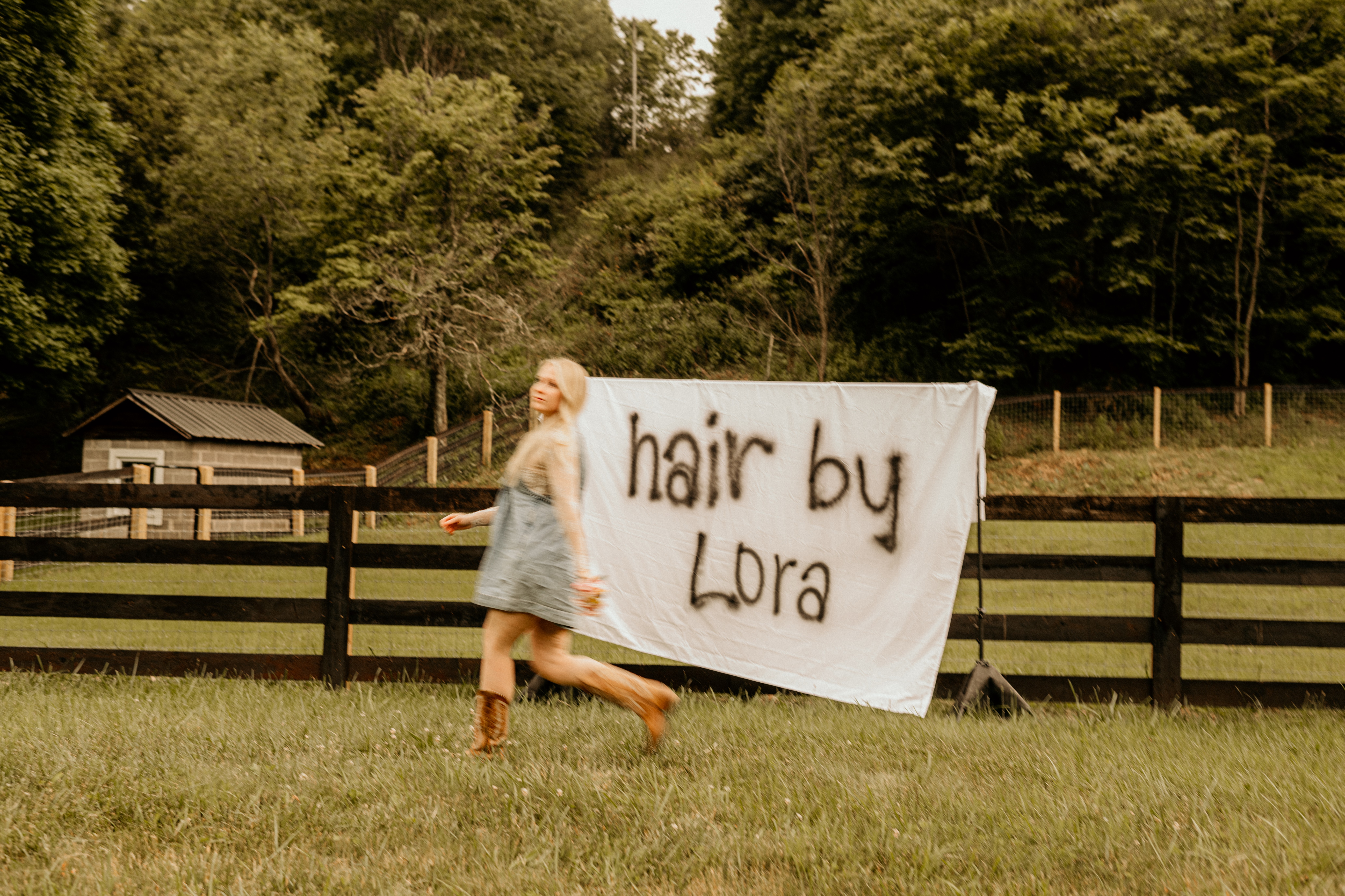 Person with light-colored hair standing on grass near a fence with a large sign reading 'hair by Lora' behind them.