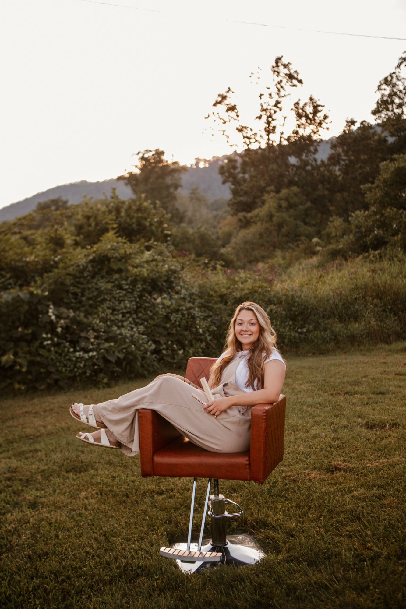 Woman sitting on a red chair outdoors with trees and hills in the background, smiling, wearing a white top and beige pants.