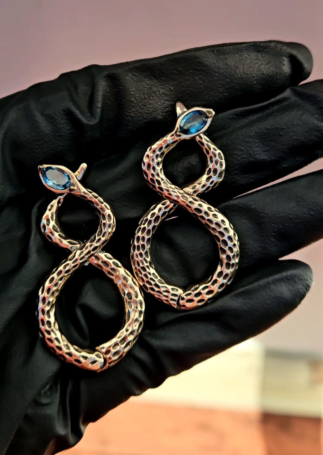 Gold snake-shaped earrings with blue gemstone accents held by a gloved hand.
