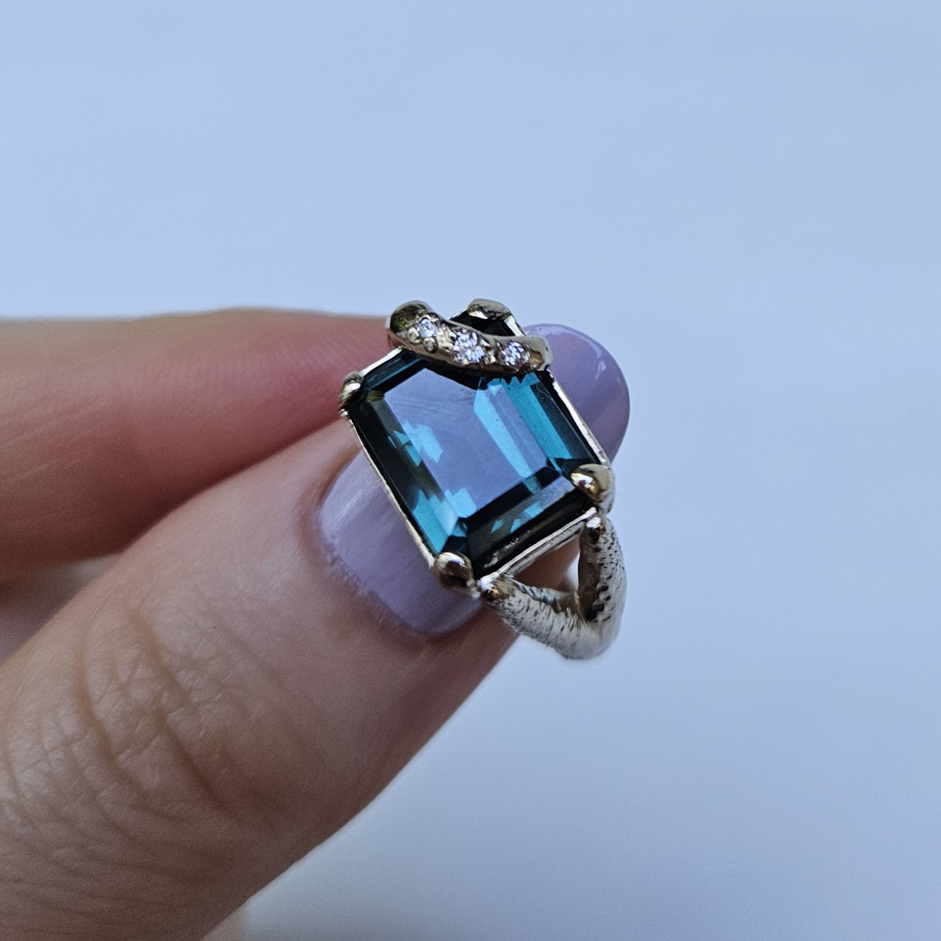 Hand holding a ring with a large blue gemstone and smaller clear stones, against a light background.