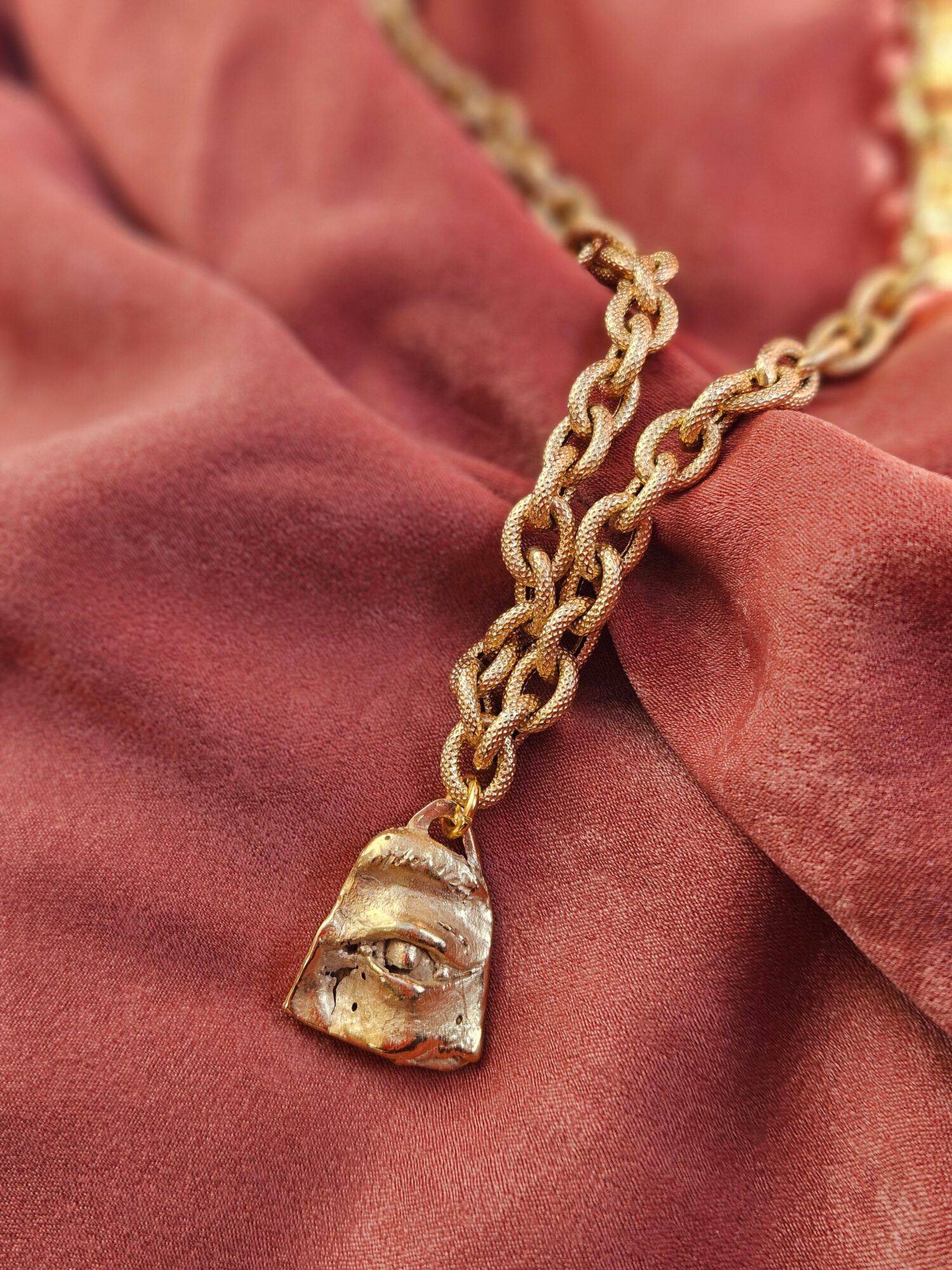 Gold chain necklace with a pendant resting on pink fabric, pendant appears to be a carved face or mask.